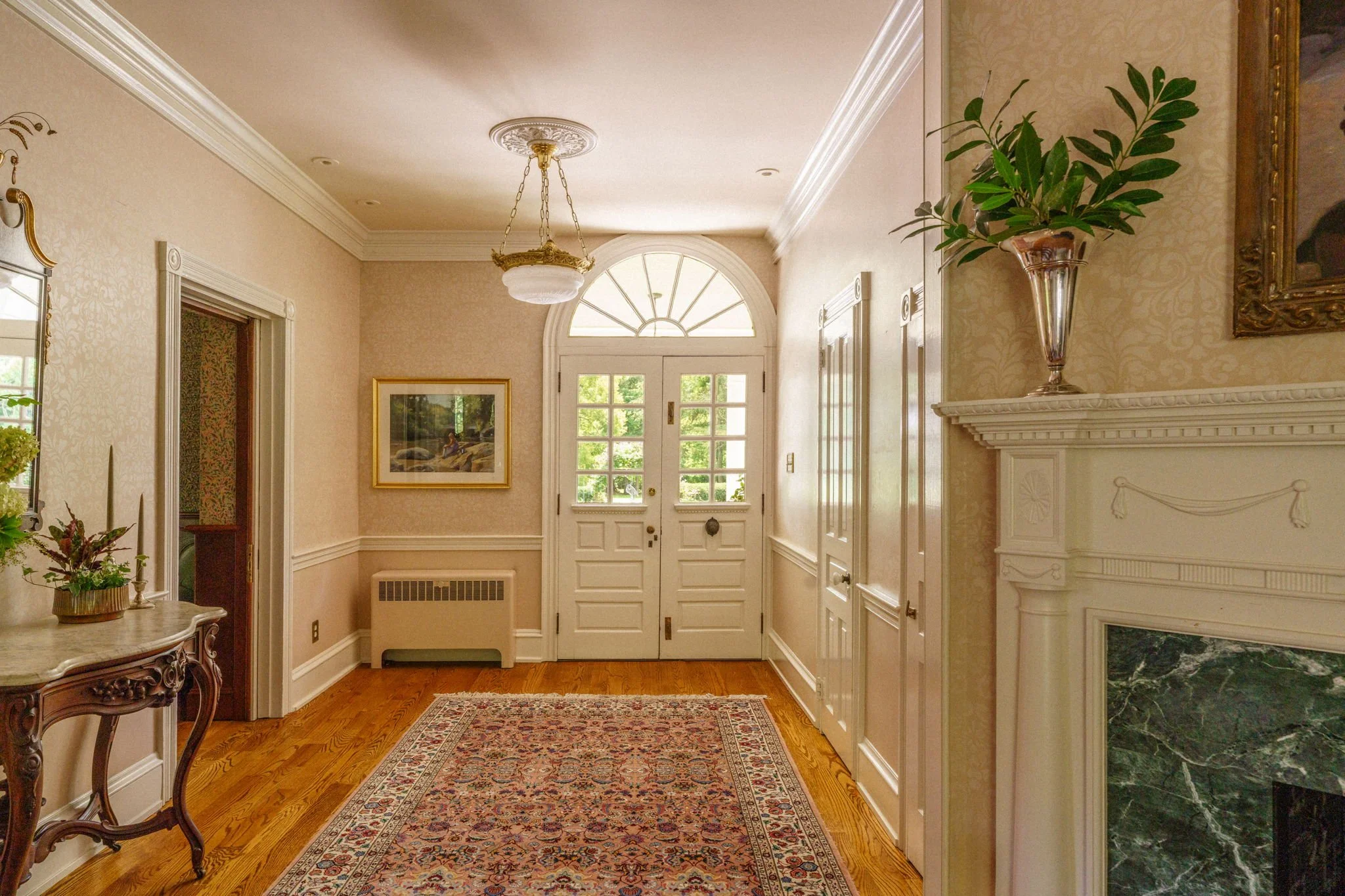 Elegant foyer with white double doors, a decorative chandelier, a framed painting on the wall, a marble-top side table with flowers, hardwood floors, and a fireplace with a large green marble hearth.