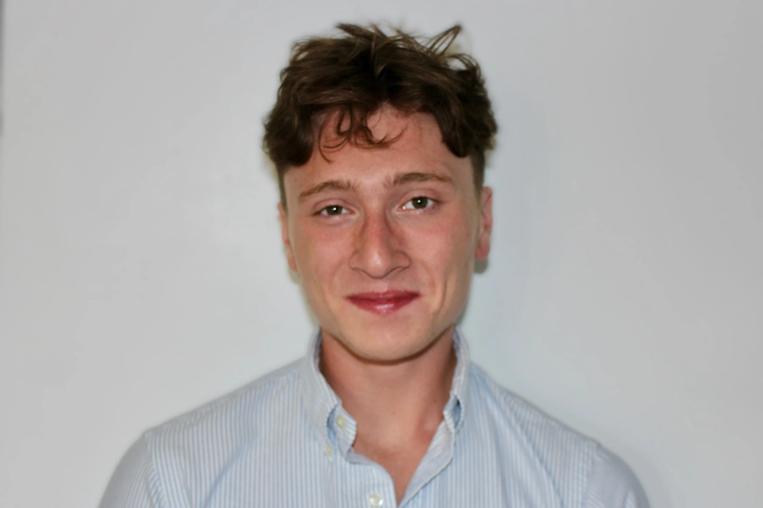 A young man with light skin, brown curly hair smiling in front of a plain light gray background, wearing a light blue collared shirt with thin white stripes.