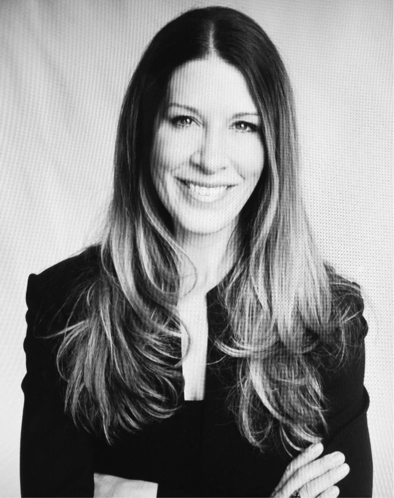 Black and white portrait of a smiling woman with long wavy hair, wearing a dark blazer, against a plain background.