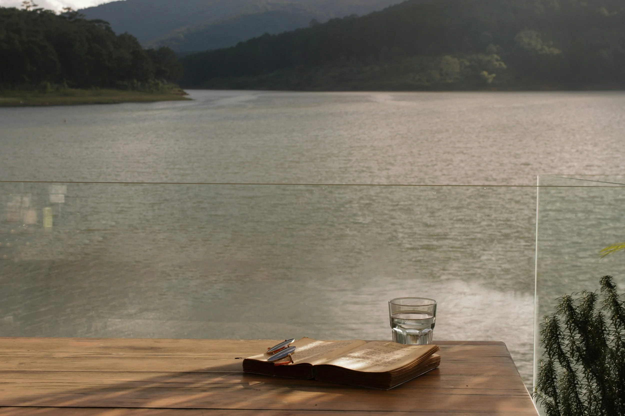 A wooden table with an open book and a glass of water, overlooking a lake with forested hills in the background.