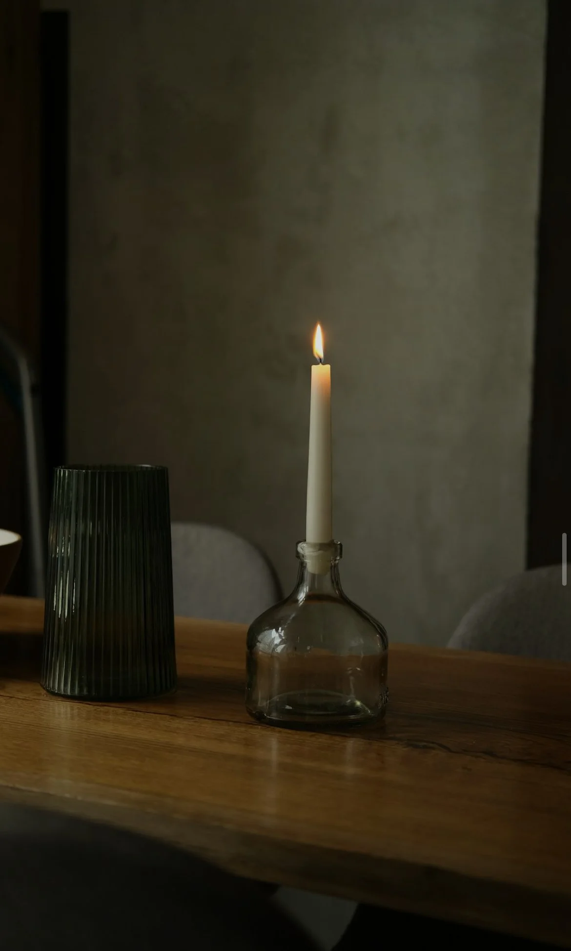 A lit white taper candle in a clear glass bottle on a wooden table, with a black ribbed vase beside it, and a beige wall in the background.