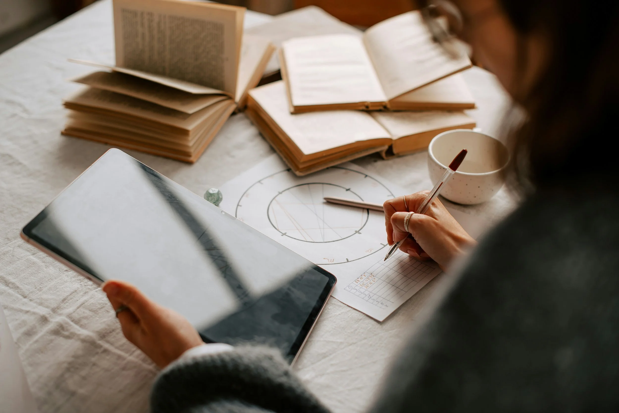 Person working at a table with open books, a tablet, a bowl, and a printed diagram with writing and geometric shapes.