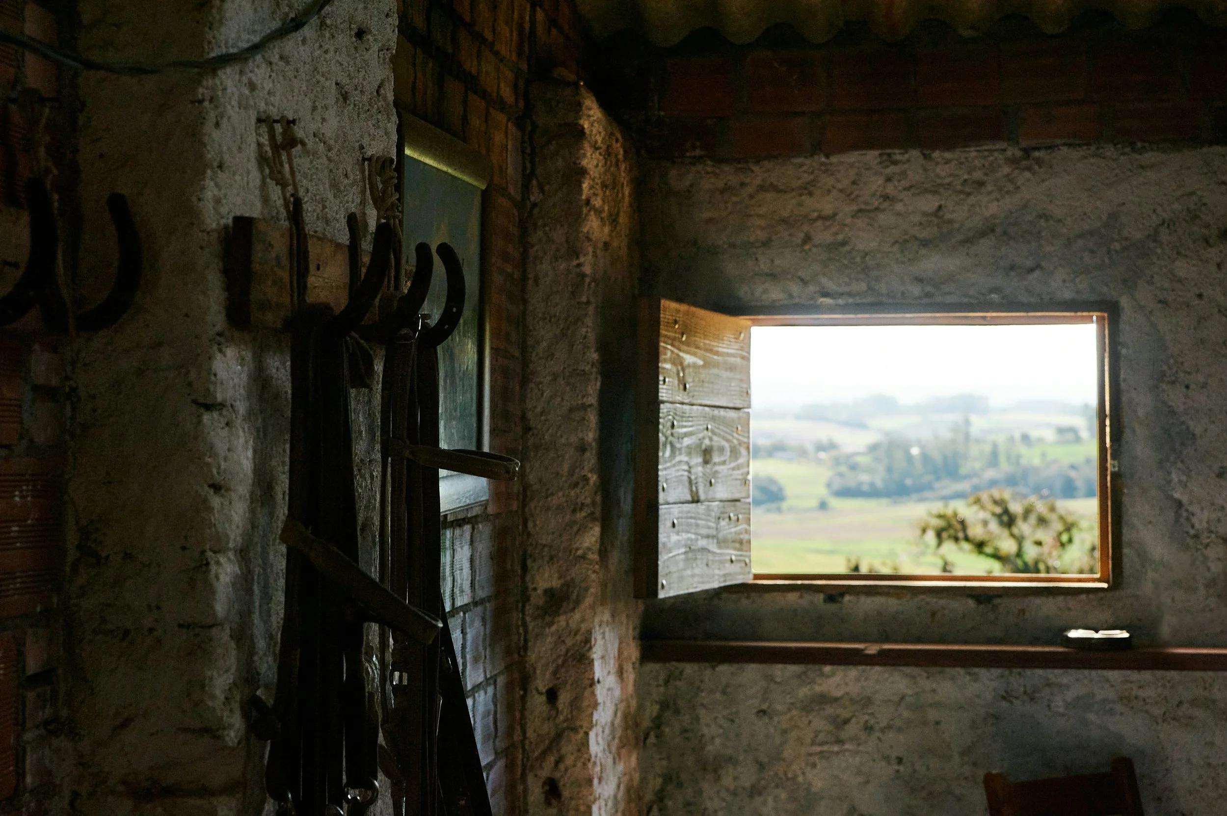 Interior view of a rustic room with a stone wall, wooden window with shutters open to a landscape, and hanging tools or equipment on hooks.