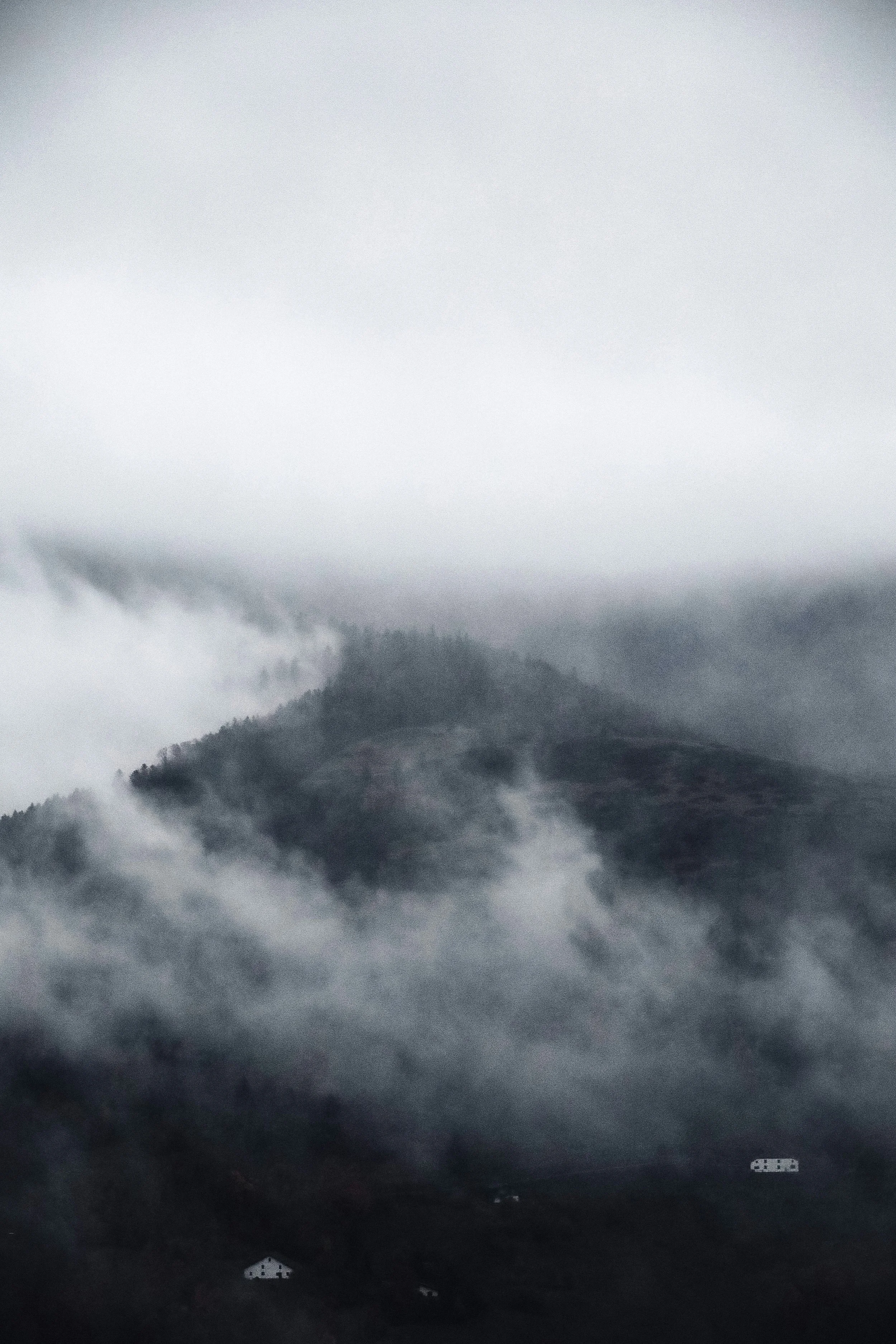 A mountain landscape shrouded in thick fog and low clouds, with a few small houses at the base.