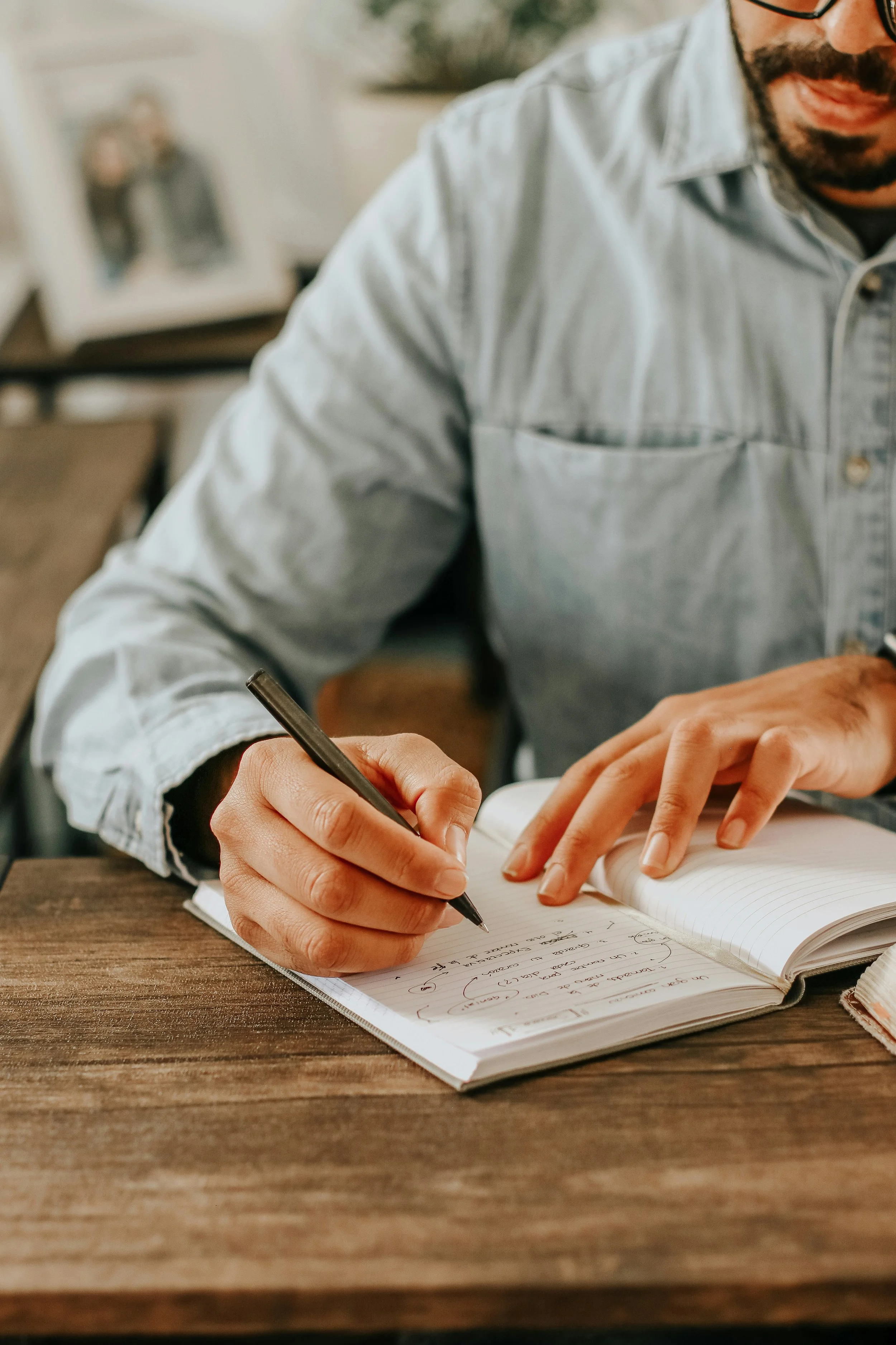 A person writing in a notebook with a black pen on a wooden table. The person is wearing a light gray denim shirt and has a beard. In the background, there is a blurred picture frame on a desk.