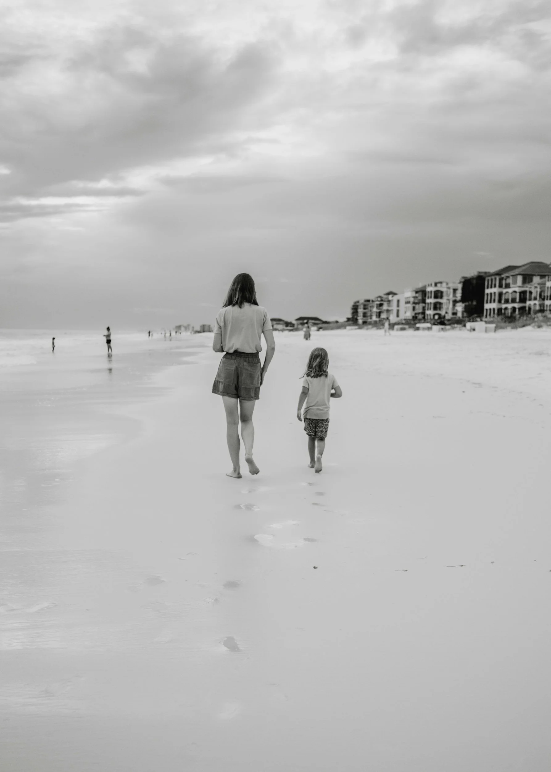 A woman and a young girl walk barefoot on the beach, leaving footprints in the wet sand, with houses in the distance under a mostly cloudy sky.