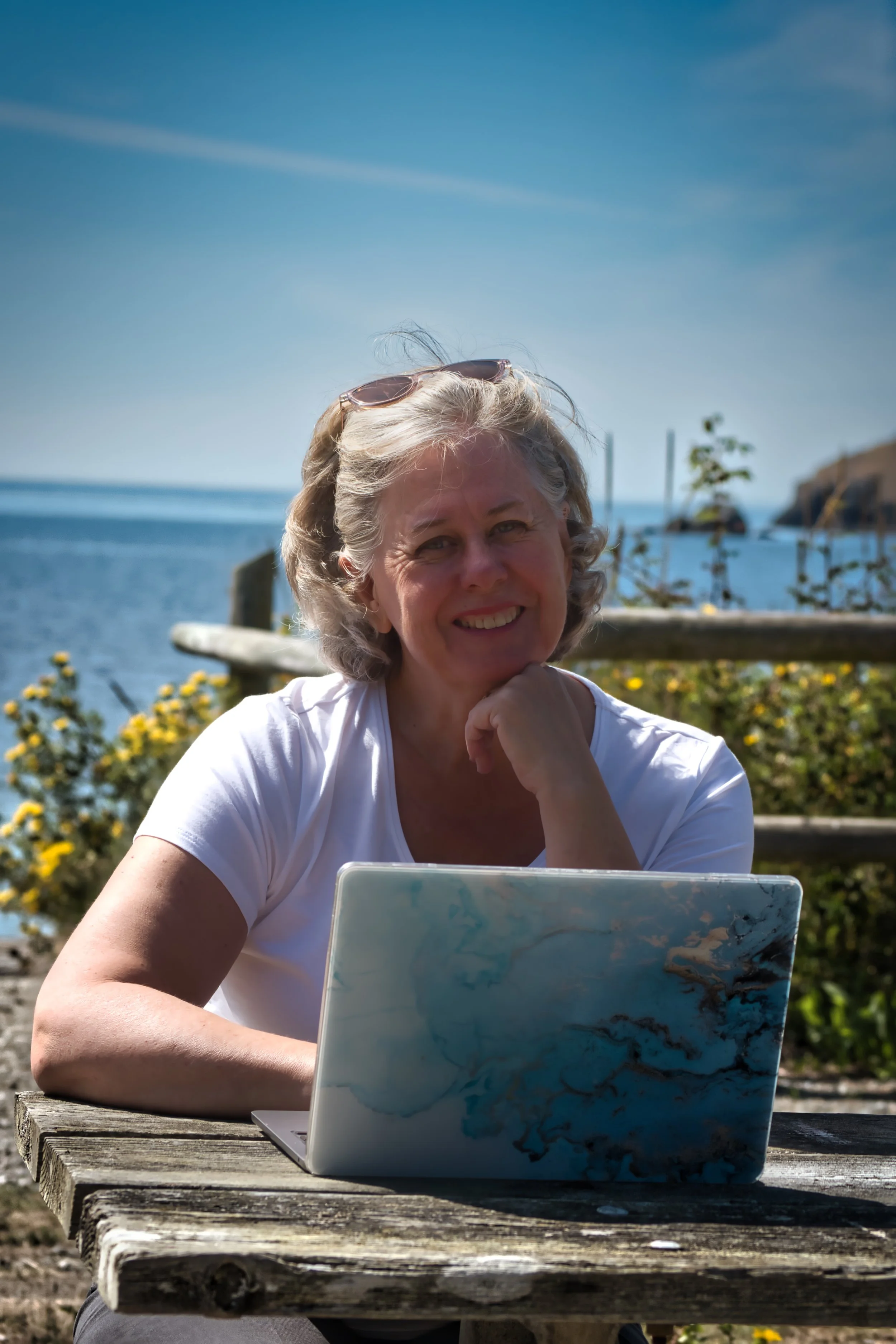 A smiling elderly woman sitting at a weathered wooden table outdoors near the ocean, with a laptop in front of her, wearing sunglasses on her head and a white t-shirt.