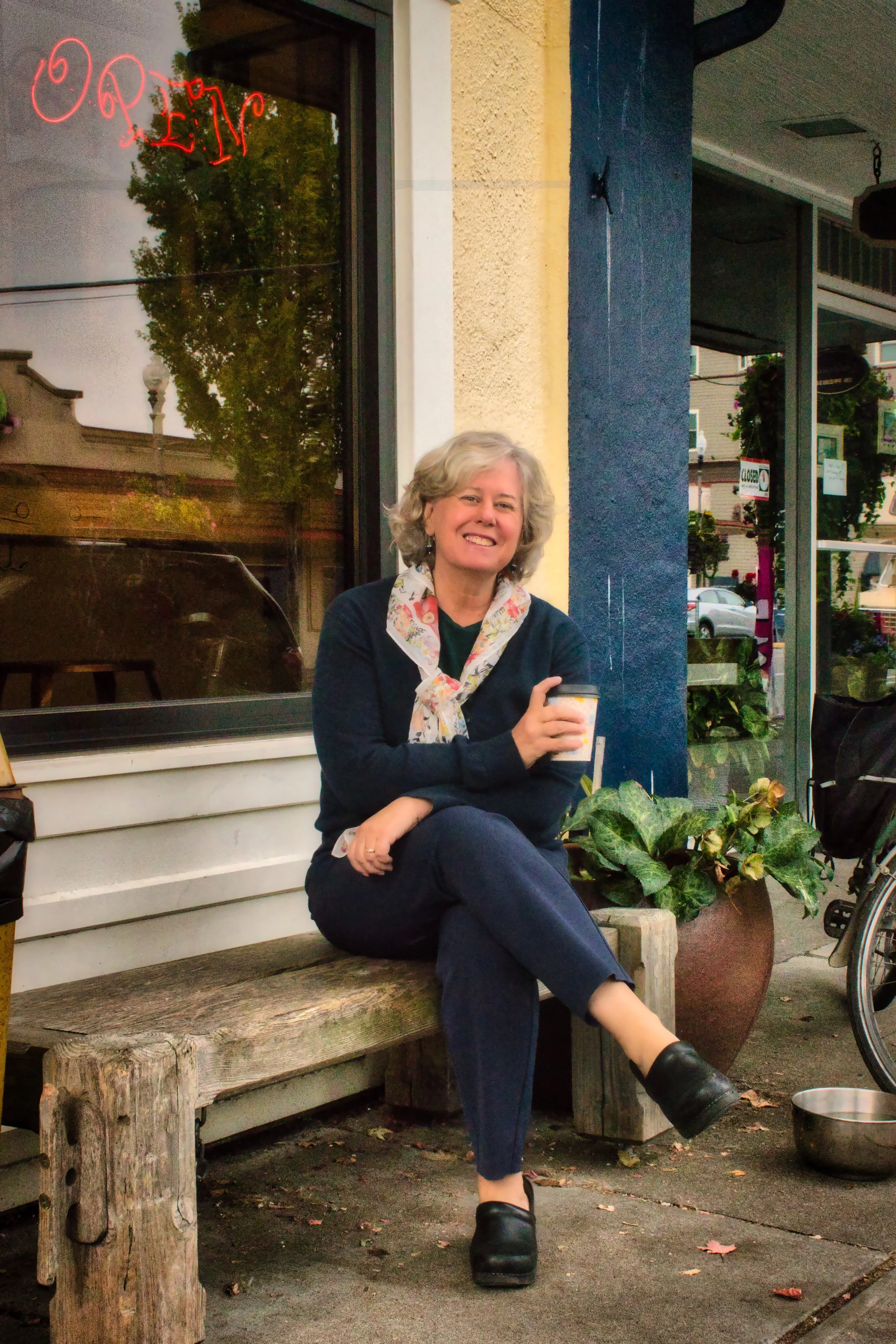 A smiling woman with curly gray hair sitting on a wooden bench outside a storefront, holding a paper cup, with a large potted plant beside her.