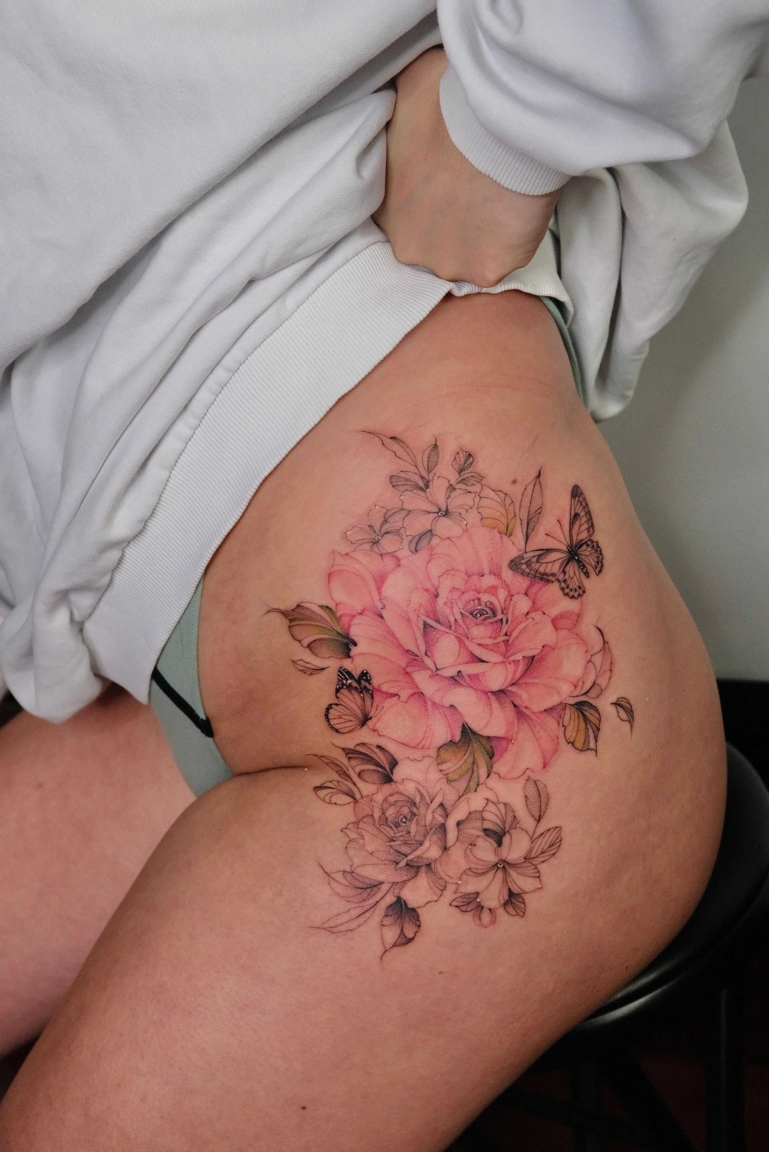 A person's thigh with a detailed tattoo of pink roses, yellow and green leaves, and black-outlined butterflies.