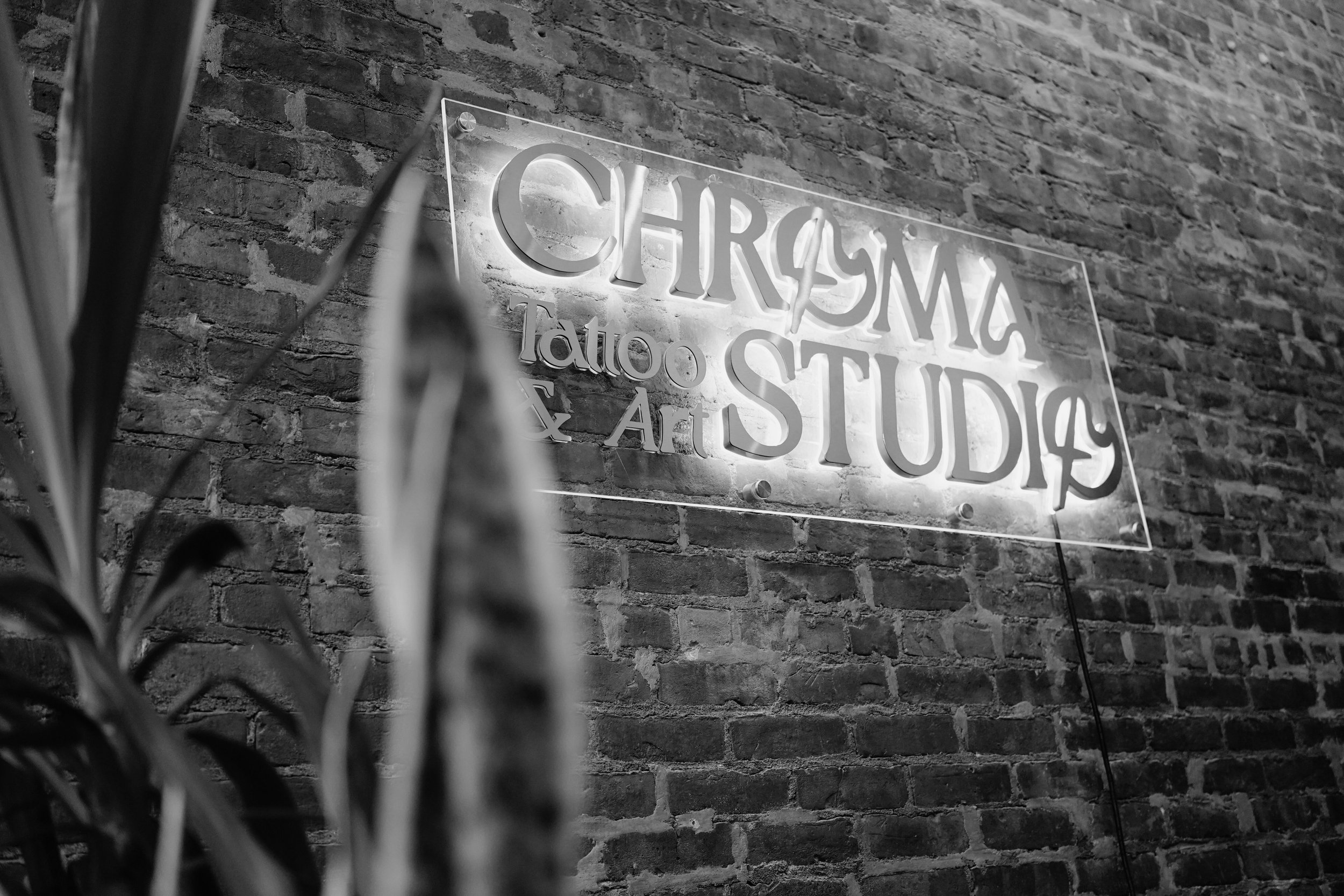 Neon sign reading 'CHROMA Tattoo & Art STUDIOS' mounted on a brick wall, with a blurred plant in the foreground.