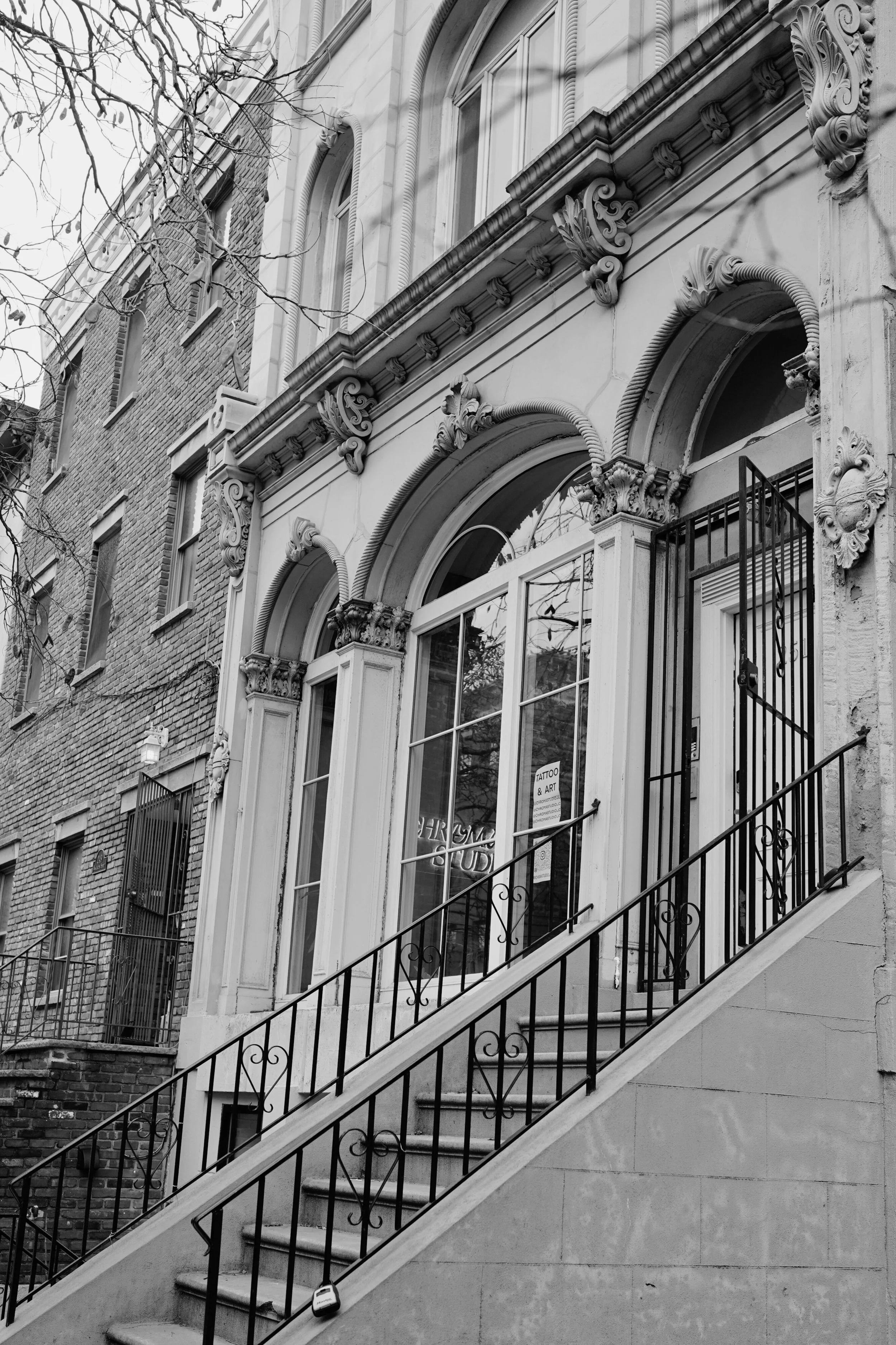 Black and white photo of a building's ornate entrance with decorative arches and columns, a staircase with an iron railing, and trees reflected in the windows.