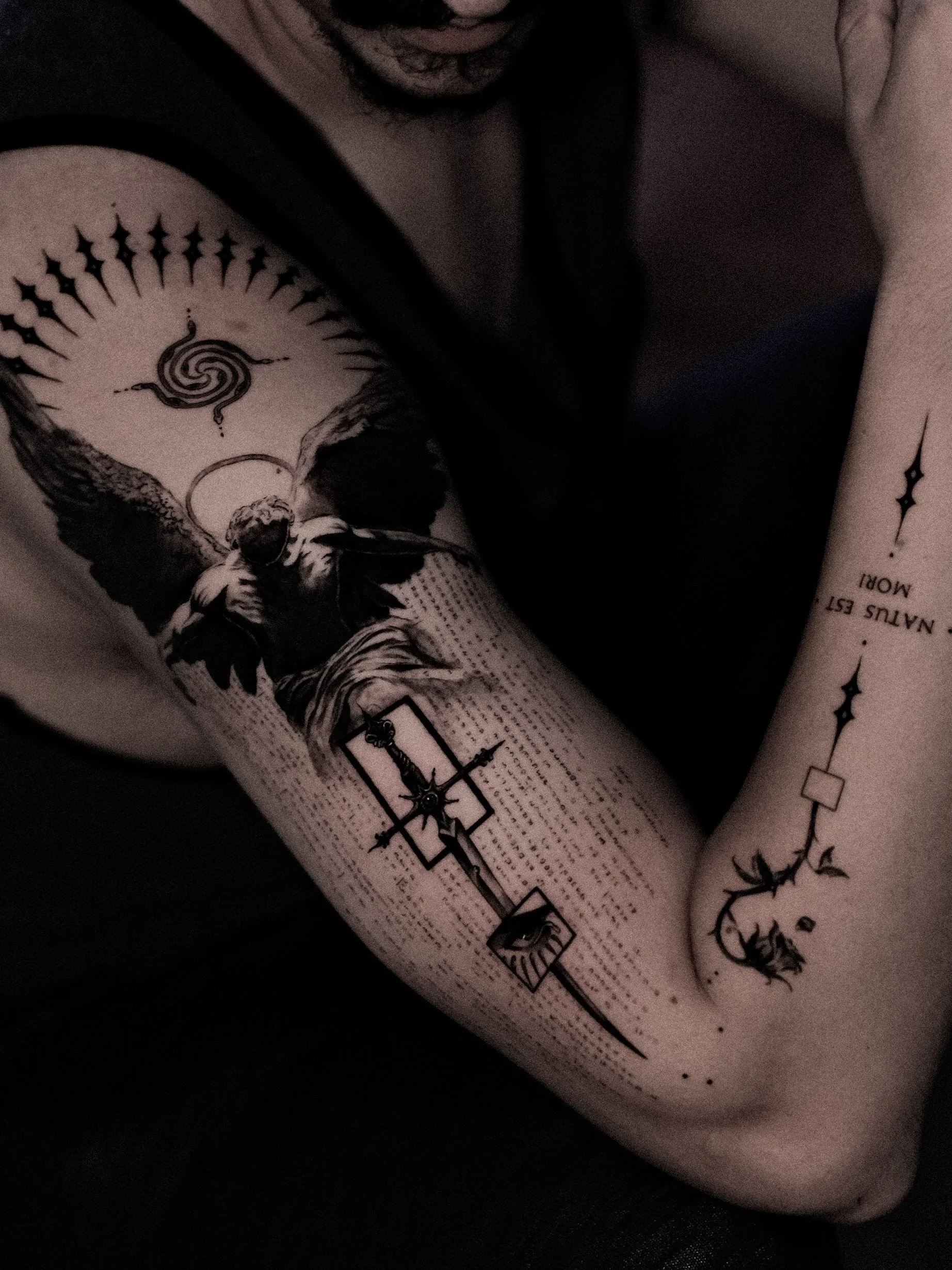 Close-up of a person's arm with black ink tattoos, including a large angel with wings and a halo, a compass with a sword, and a line of text with a star and floral design elements.