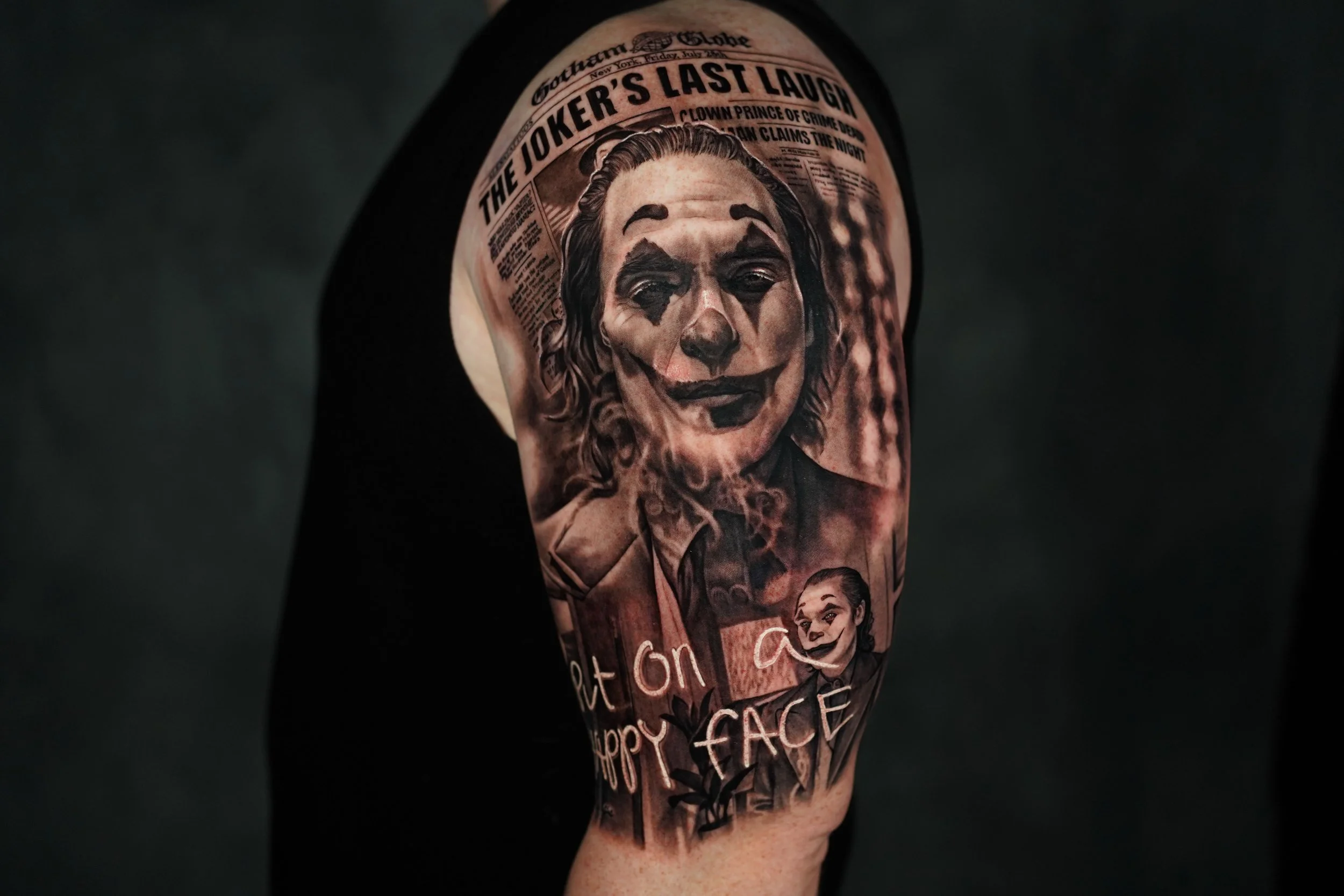 Black and white tattoo of the Joker from the movie 'The Joker's Last Laugh' on a person's upper arm, featuring a clown face with makeup and a newspaper headline.