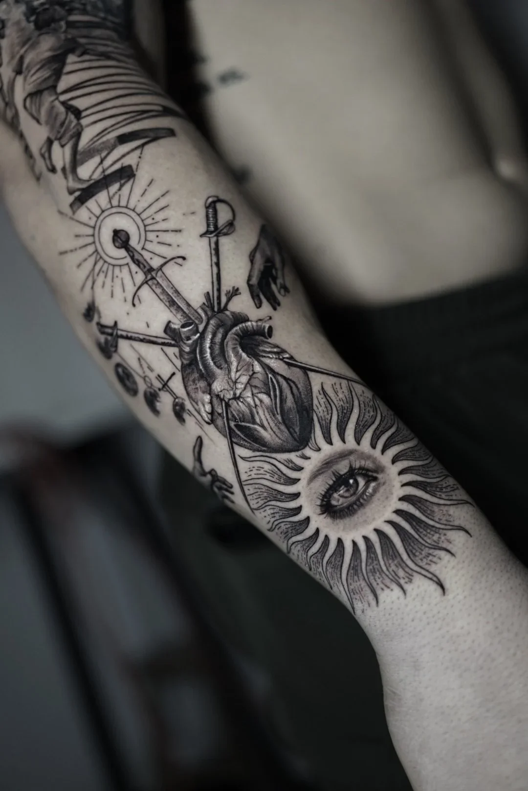 Tattoo of a realistic heart with a sword, sun with an eye, and other elements in black and white ink.