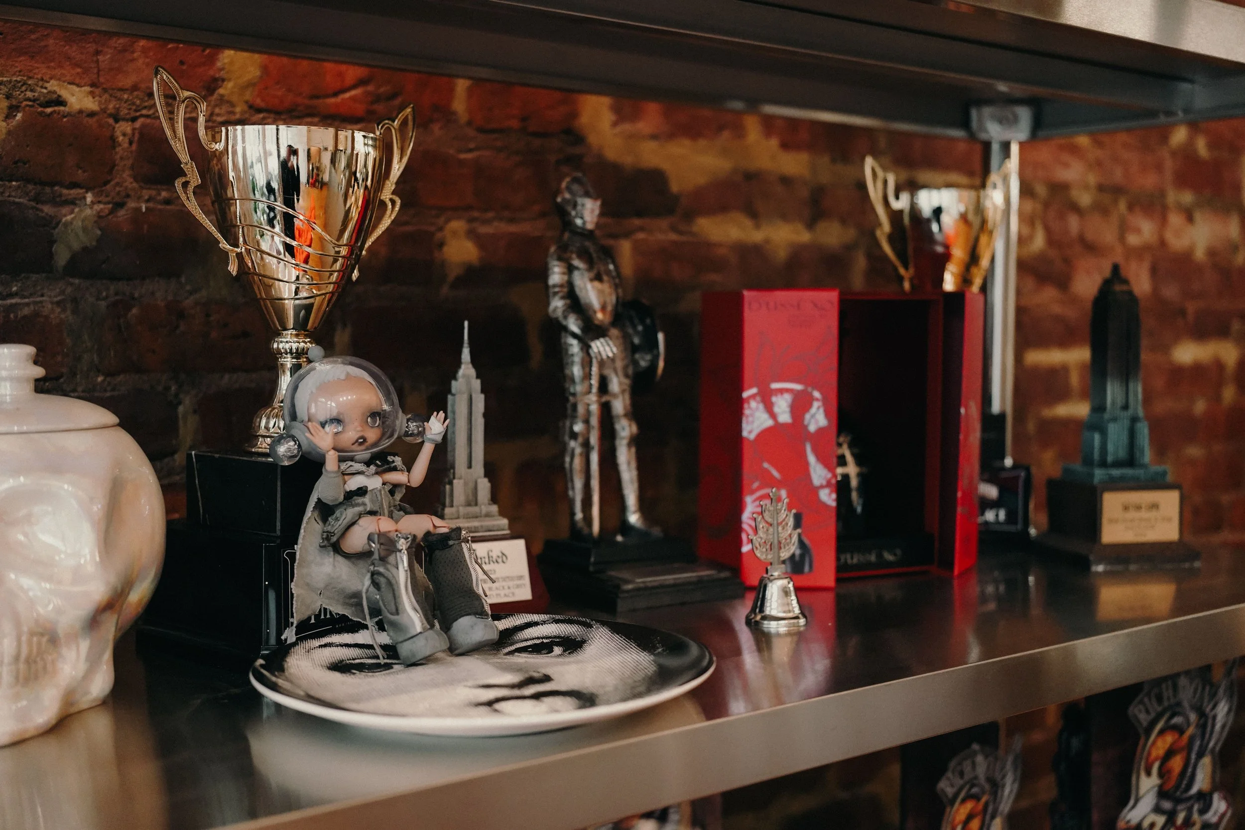 A collection of decorative items on a metal shelf against a brick wall, including a doll, trophies, a miniature statue, and various books or boxes.