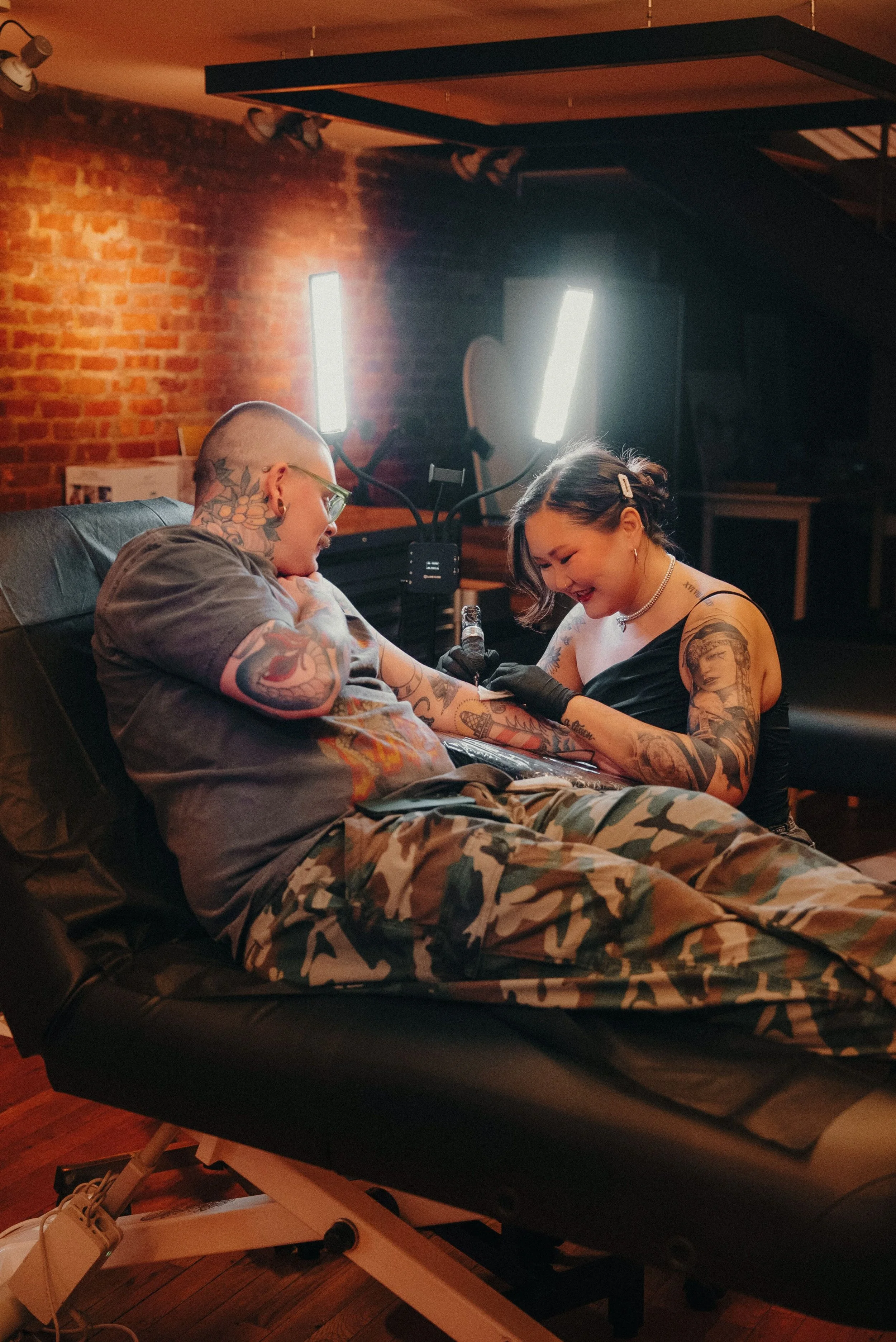 A woman with tattoos getting a tattoo on her arm by a tattoo artist in a studio with brick walls and bright lights.