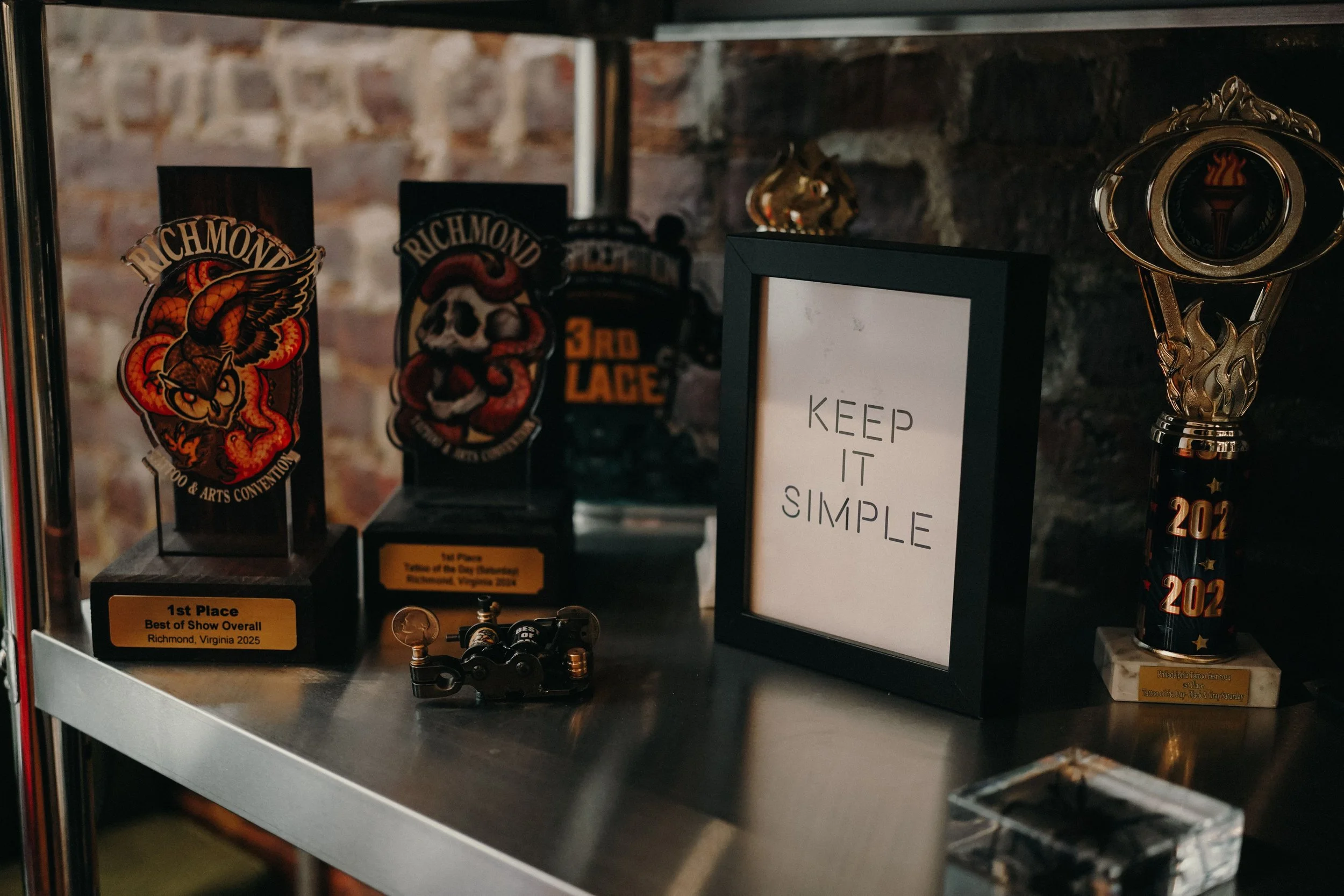 Display of trophies and awards on a metal shelf, including plaques for Richmond tattoo conventions, a framed quote that says 'Keep It Simple,' and a small model of a motorcycle, all set against a brick wall.