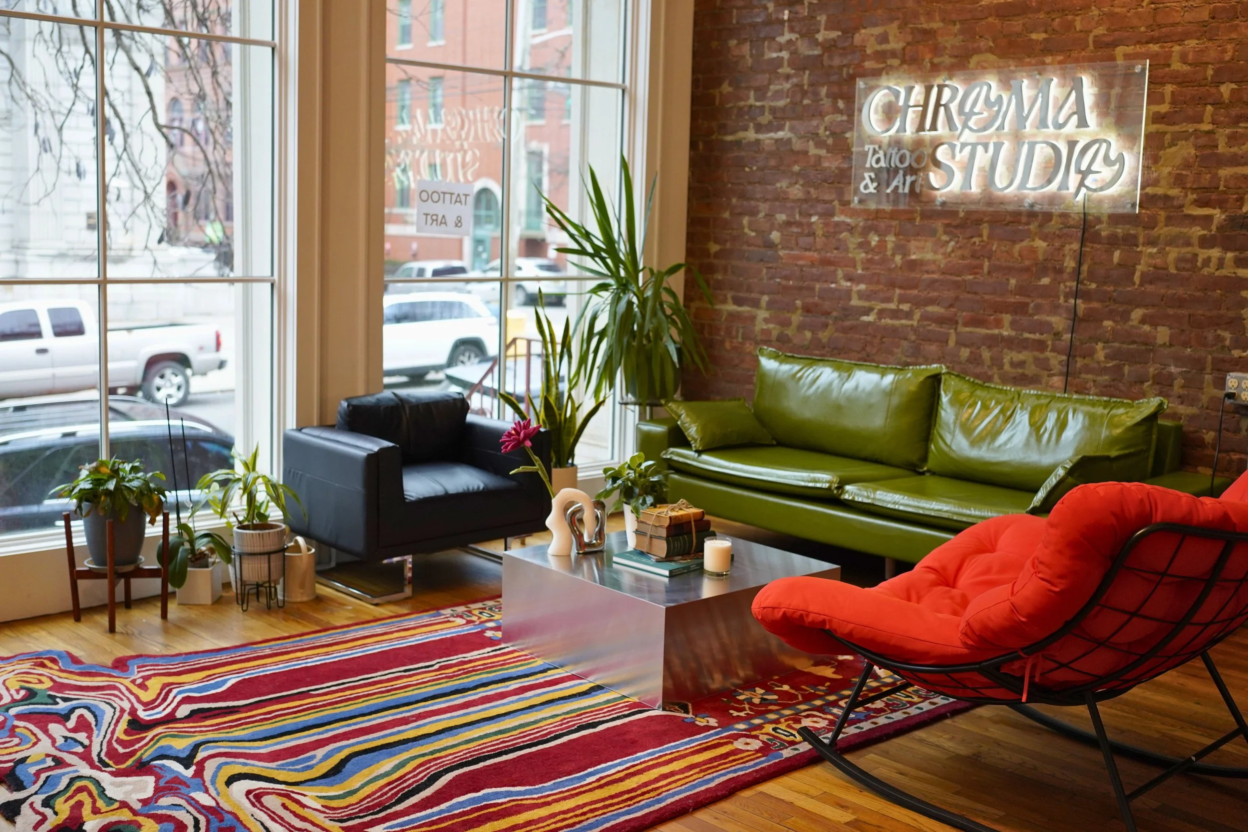 A cozy interior of an art studio with a large window, a red brick wall, and neon sign that reads 'Chroma Tattoo & Art Studio'. There are various green plants, a black armchair, a green leather couch, a red lounge chair, a colorful patterned rug, and 