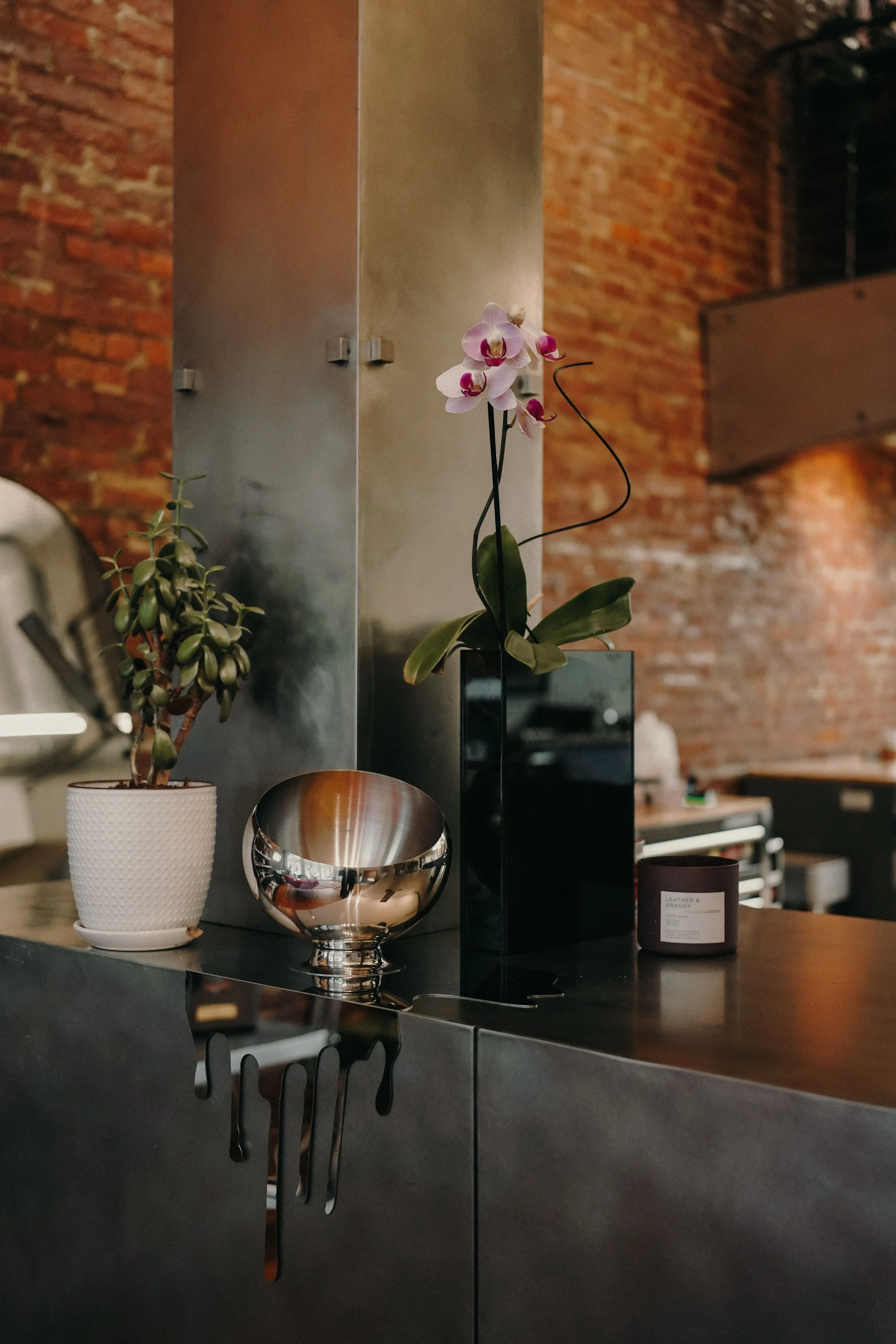 A modern kitchen or living space with a brick wall, metal cabinet, and a dark wooden surface. On the surface, there is a potted jade plant, a reflective silver bowl, a black vase with pink orchids, and a brown candle.