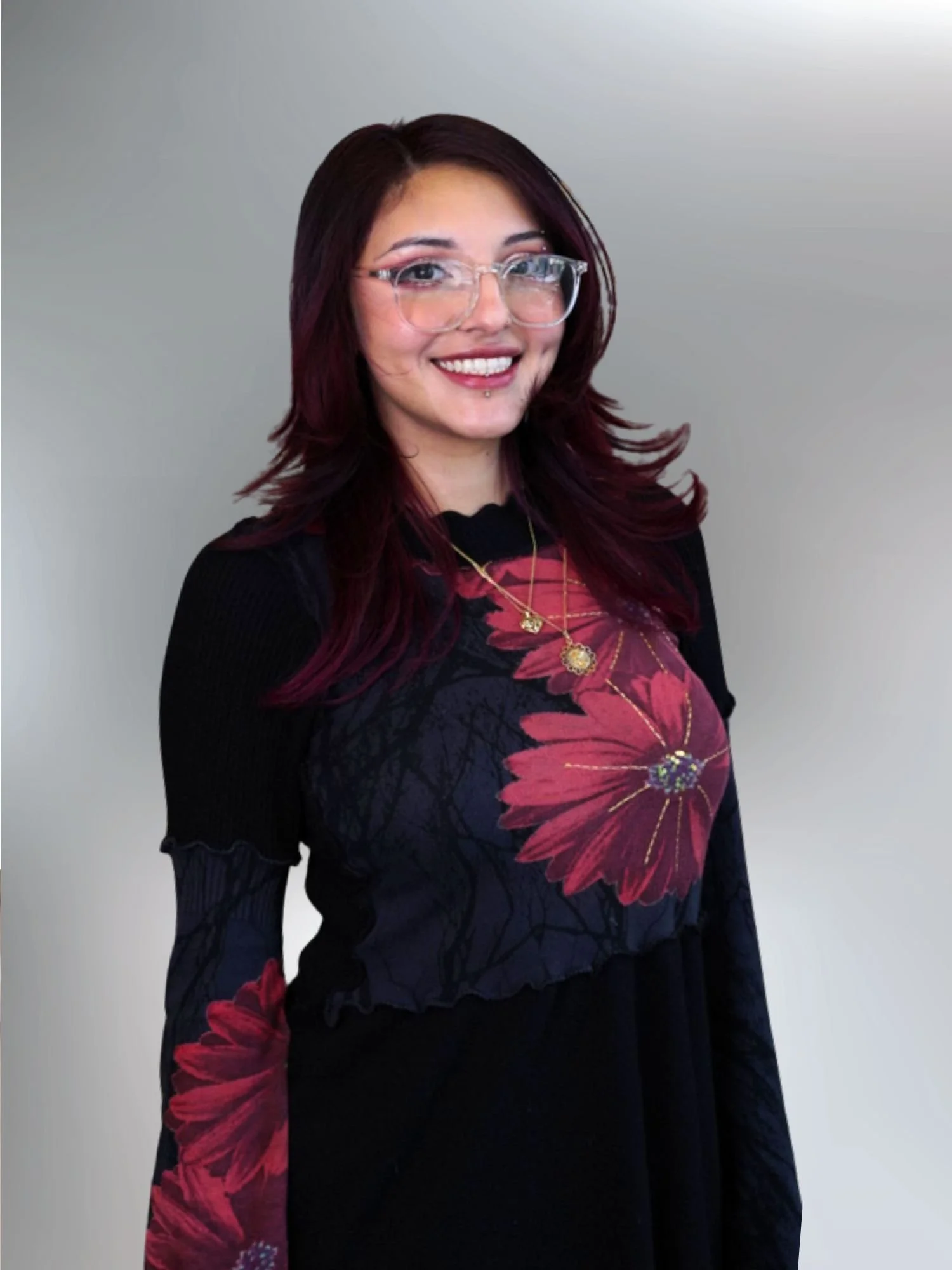 A woman with dark red hair and glasses smiling, wearing a black dress with red floral patterns and layered gold necklaces. She is standing against a plain, light-colored background.