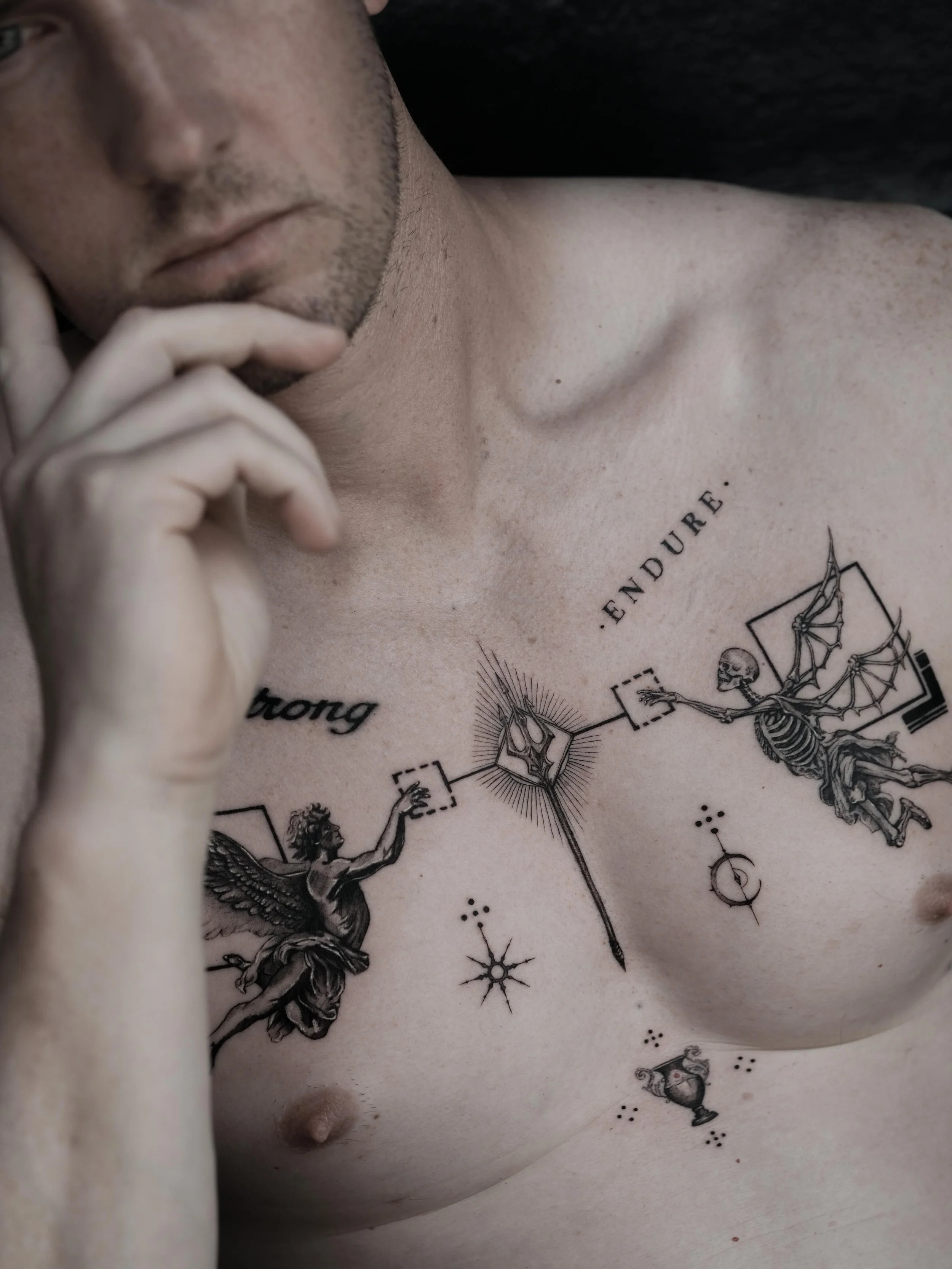 Close-up of a man's chest with black ink tattoo featuring mythological and celestial symbols, including the words "ENDURE" and "STRONG".