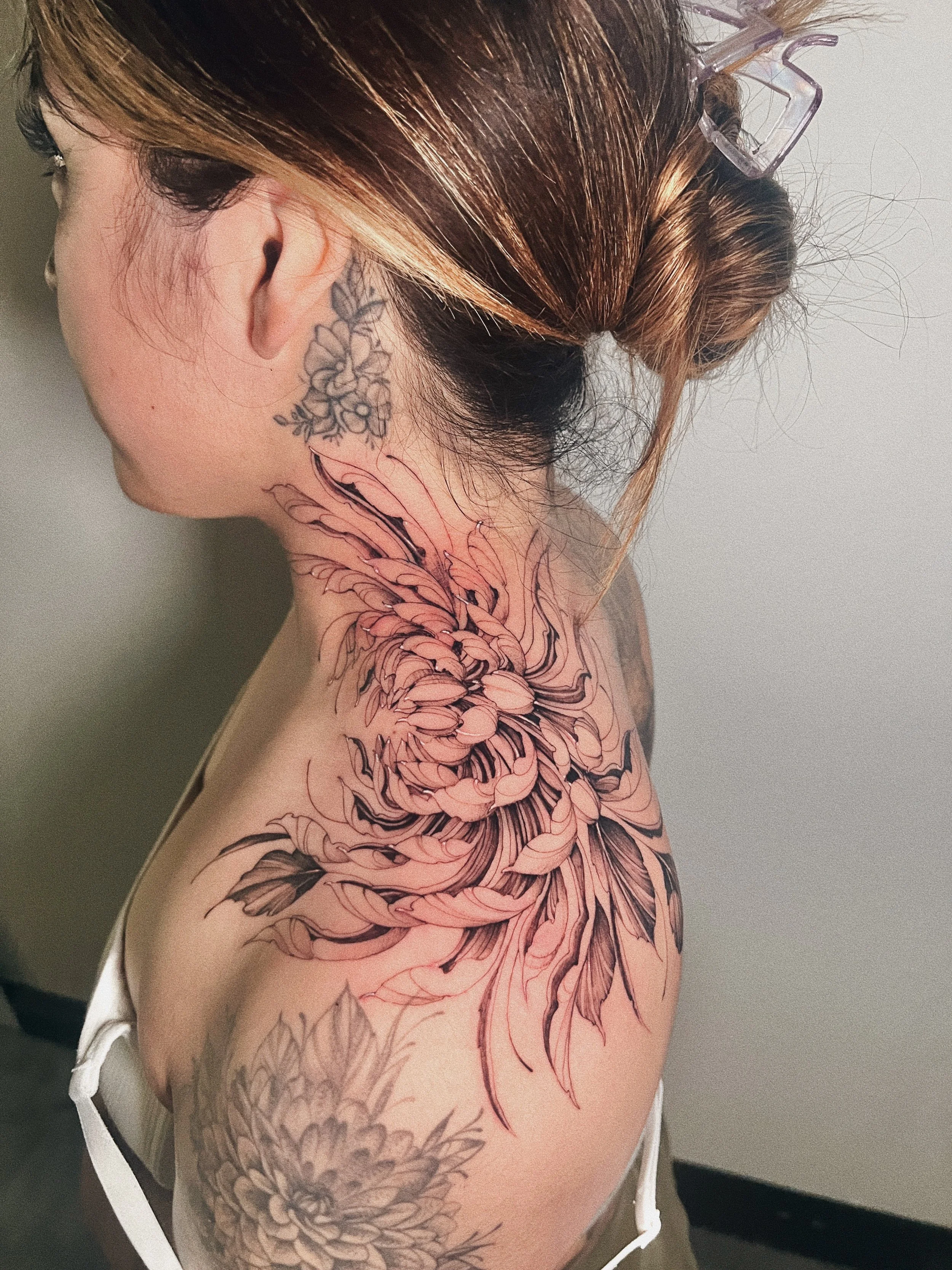 A woman with brown hair in a bun wearing glasses, showing tattoos on her neck and shoulder. The tattoos include a floral design on her neck and a large, detailed floral tattoo extending onto her shoulder.