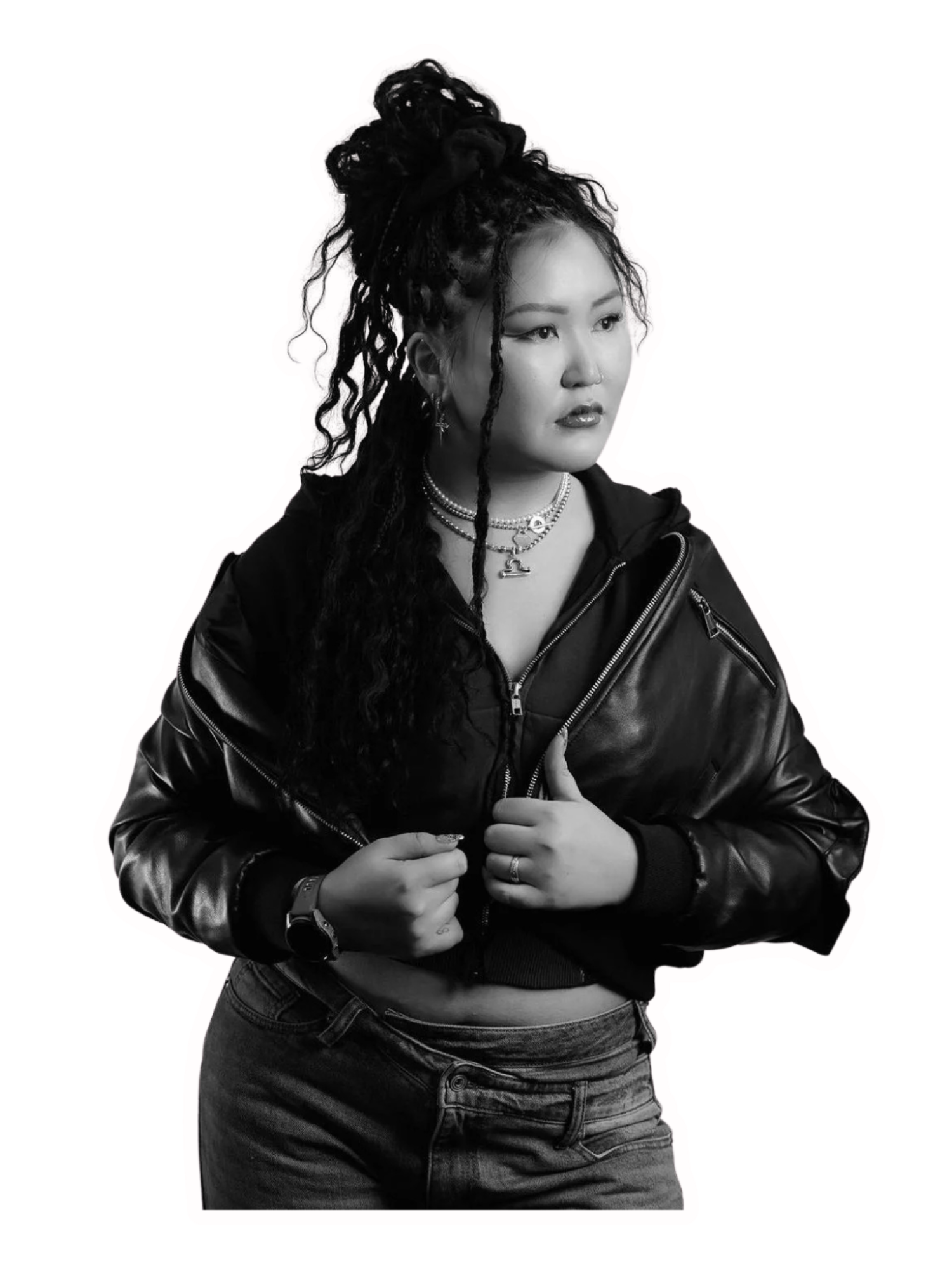 Black and white photo of a young woman with curly hair tied up in a high bun, wearing a leather jacket, necklaces, and jeans, posing confidently.