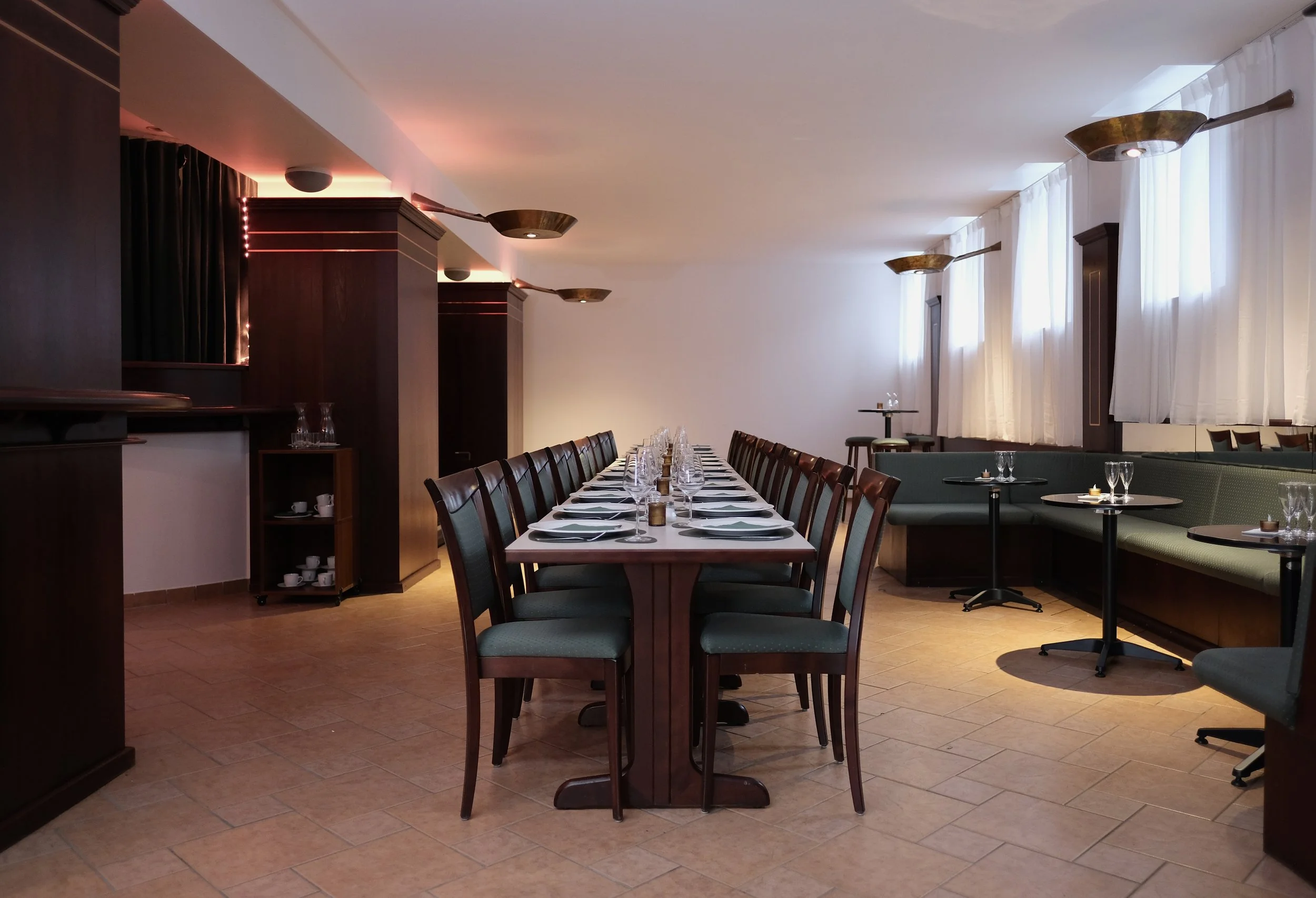 Empty restaurant with a long dining table set with plates, silverware, and glassware, surrounded by chairs, and additional small tables with glasses and napkins in the background.
