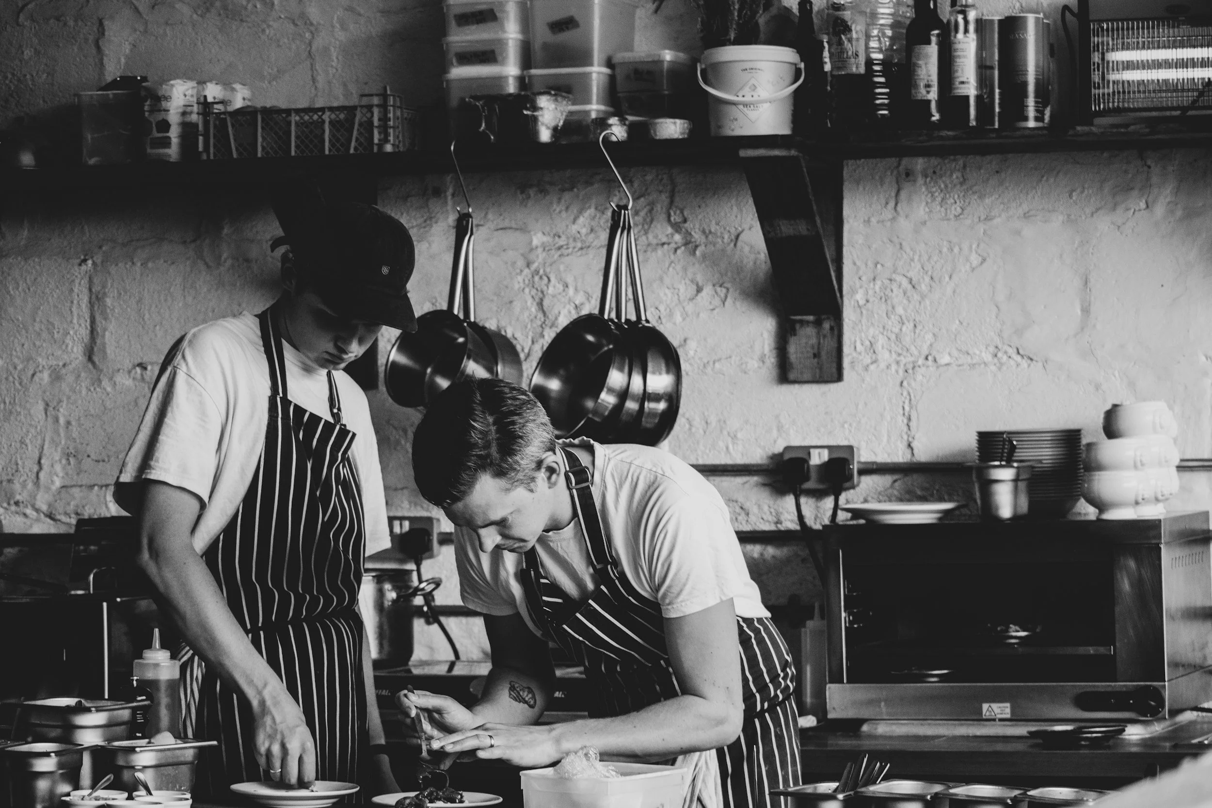 Two chefs in striped aprons preparing food in a professional kitchen, one pouring ingredients and the other focusing on a task, with pots hanging on a textured wall background.
