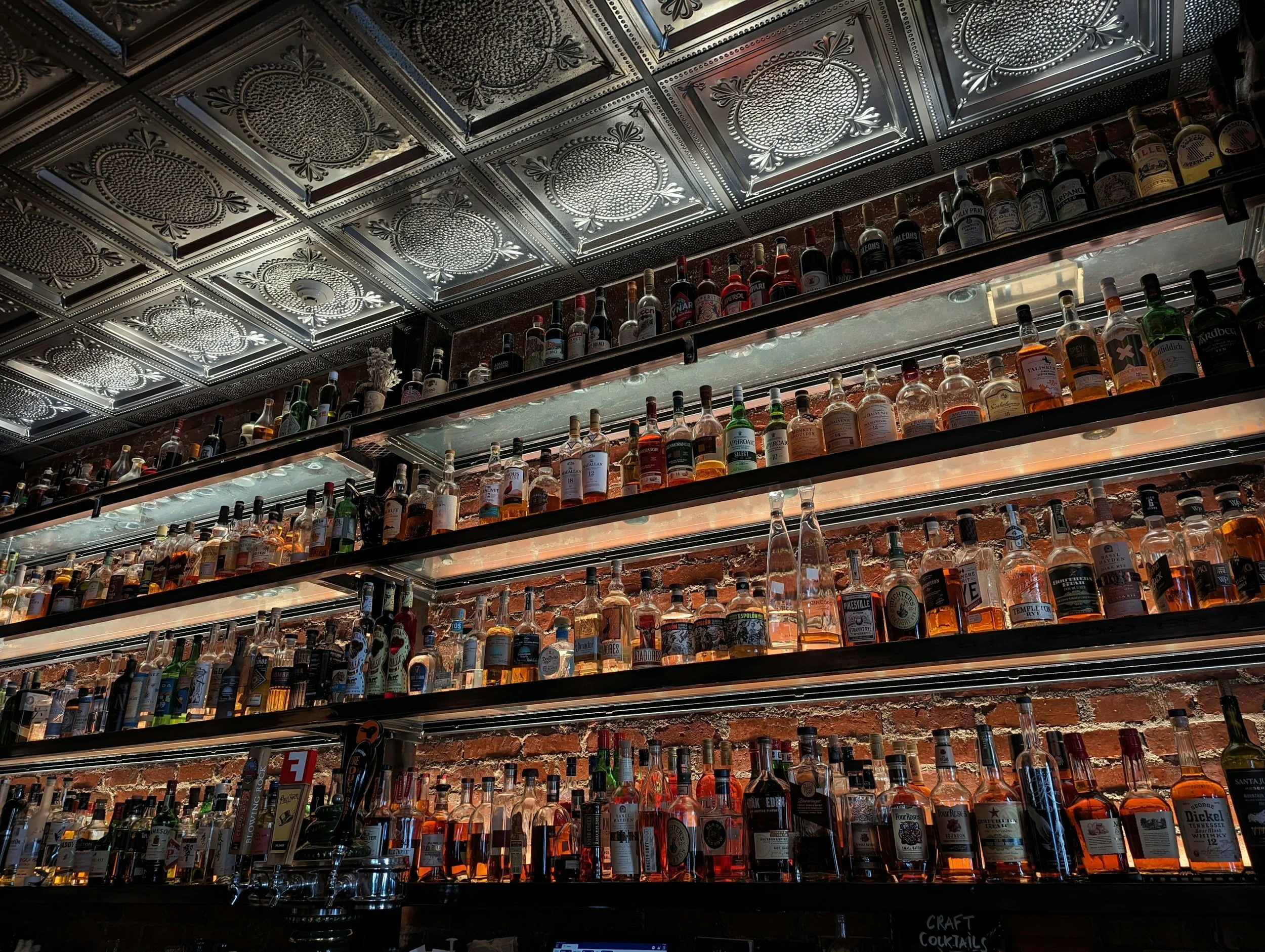 Multiple shelves filled with various bottles of liquor in a bar, against a brick wall, with a decorative metal ceiling above.