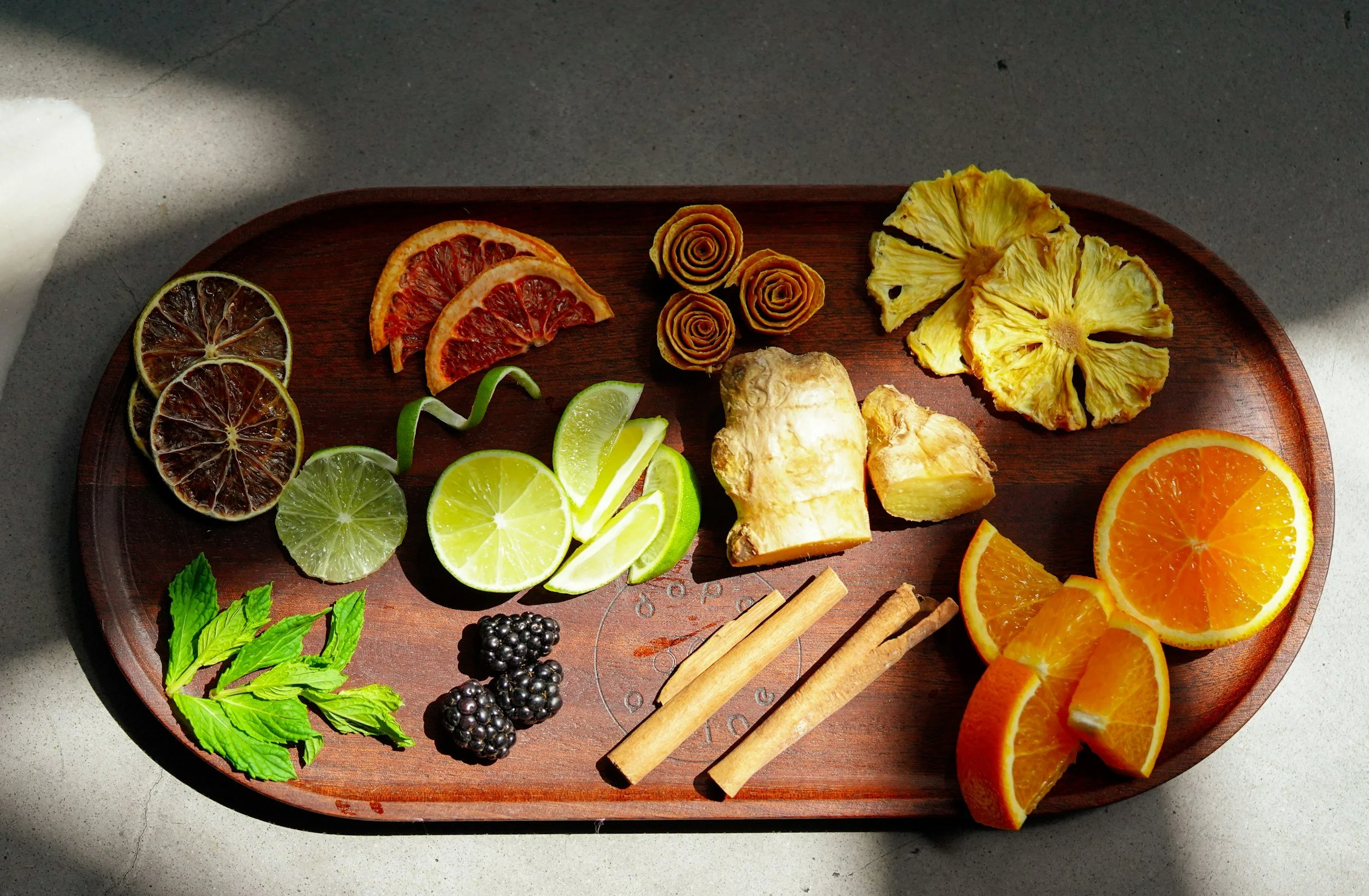 A wooden tray with dried citrus slices, cinnamon sticks, fresh blackberries, mint leaves, and sliced lime, lemon, orange, and pineapple.
