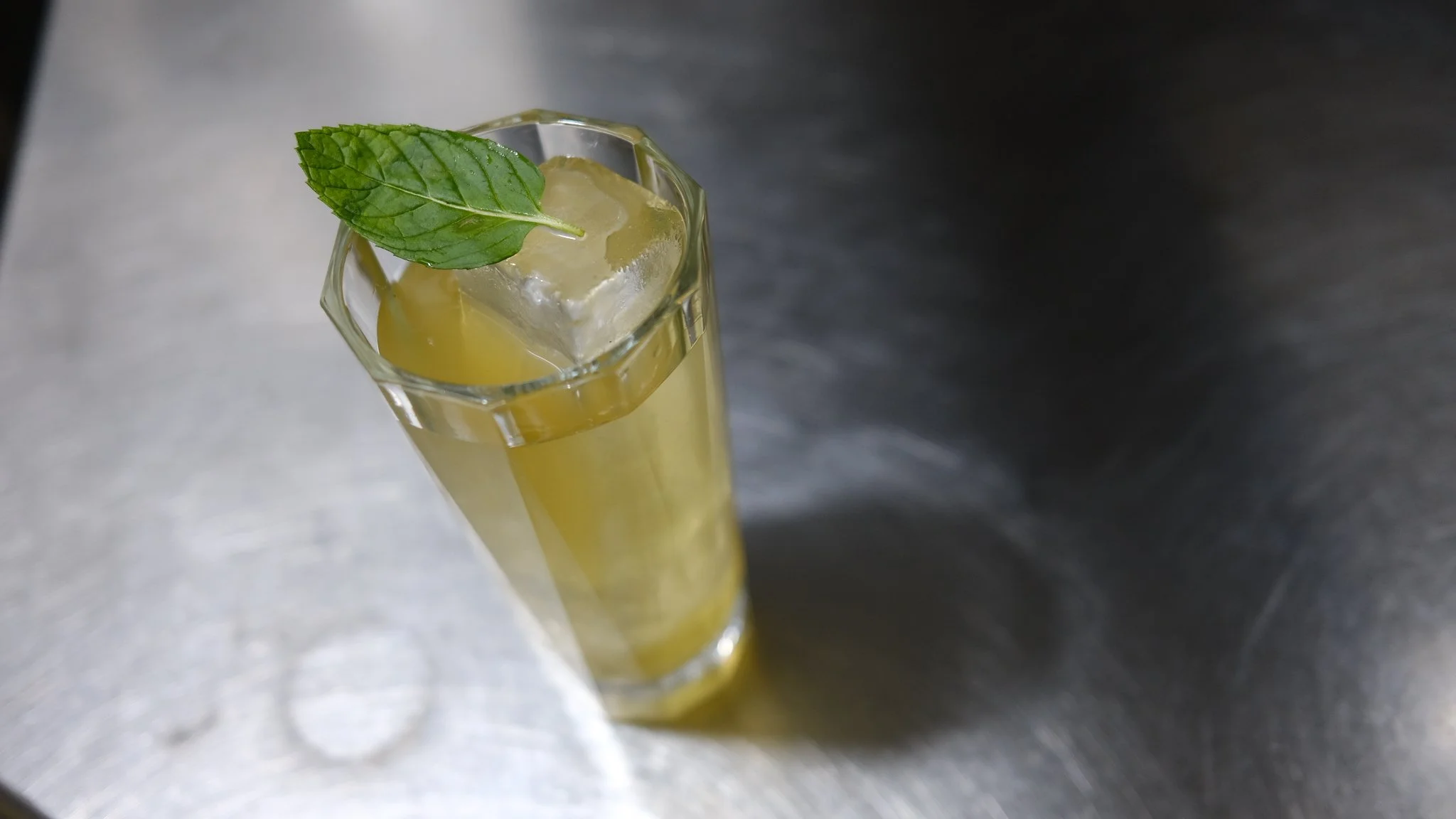 A tall glass with a yellow drink, ice cubes, and a mint leaf on top, placed on a metallic surface.