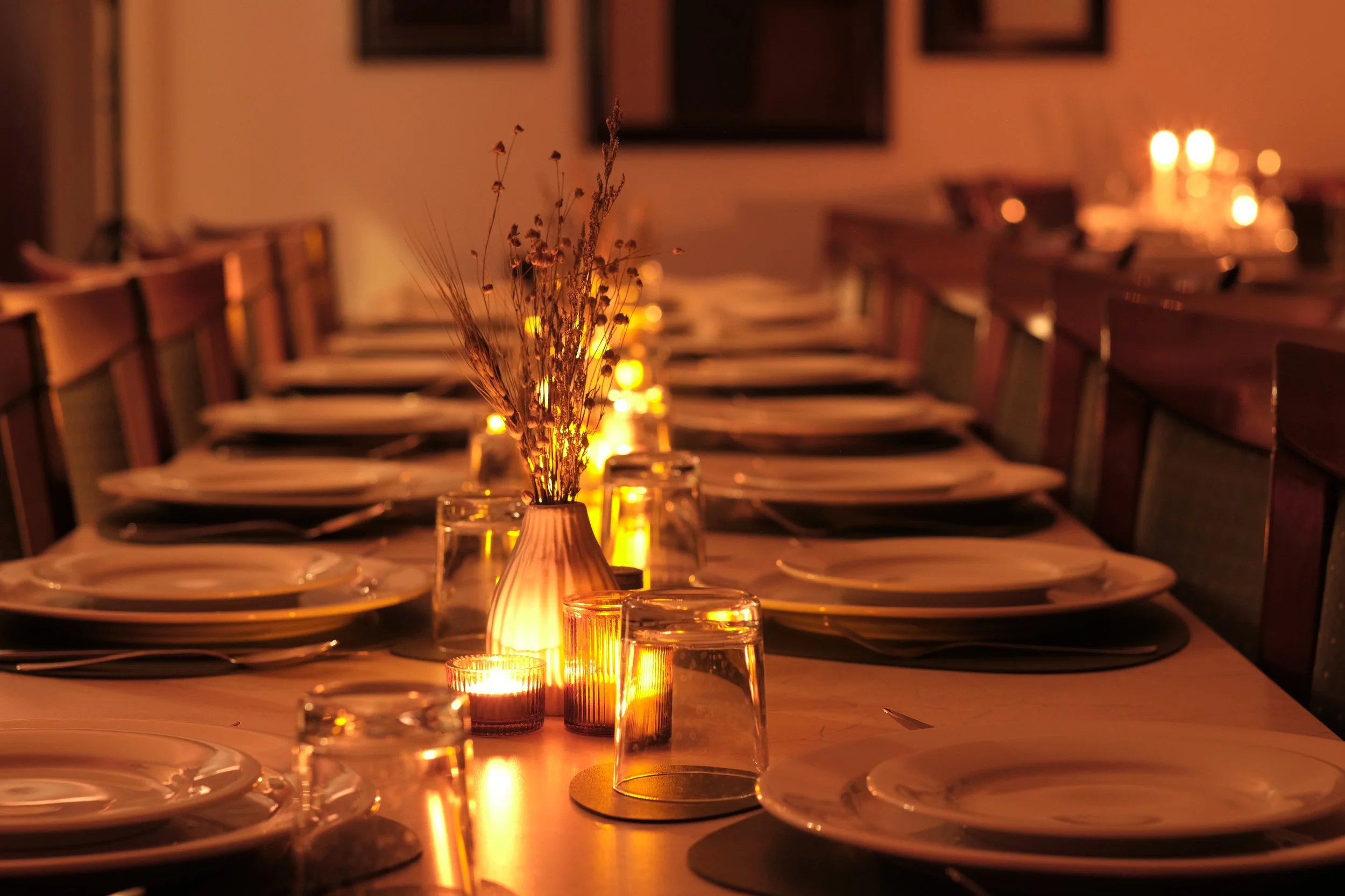 A long dining table set for a special occasion with plates, glasses, candles, and a vase of dried flowers, illuminated by warm candlelight in a cozy, dimly lit room.
