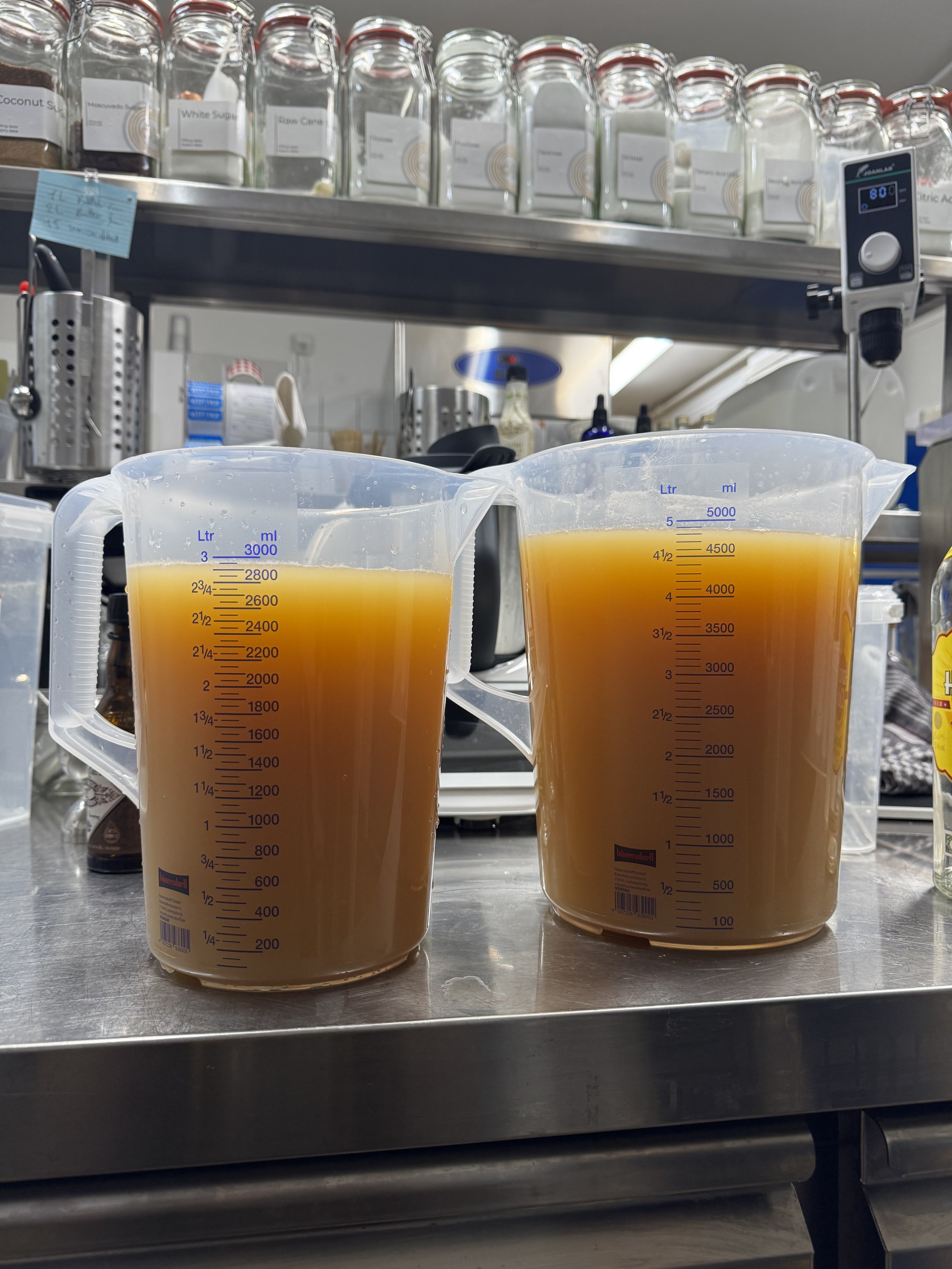 Two measuring pitchers filled with a light brown liquid, placed on a stainless steel countertop in a kitchen or laboratory setting with jars, bottles, and kitchen tools in the background.
