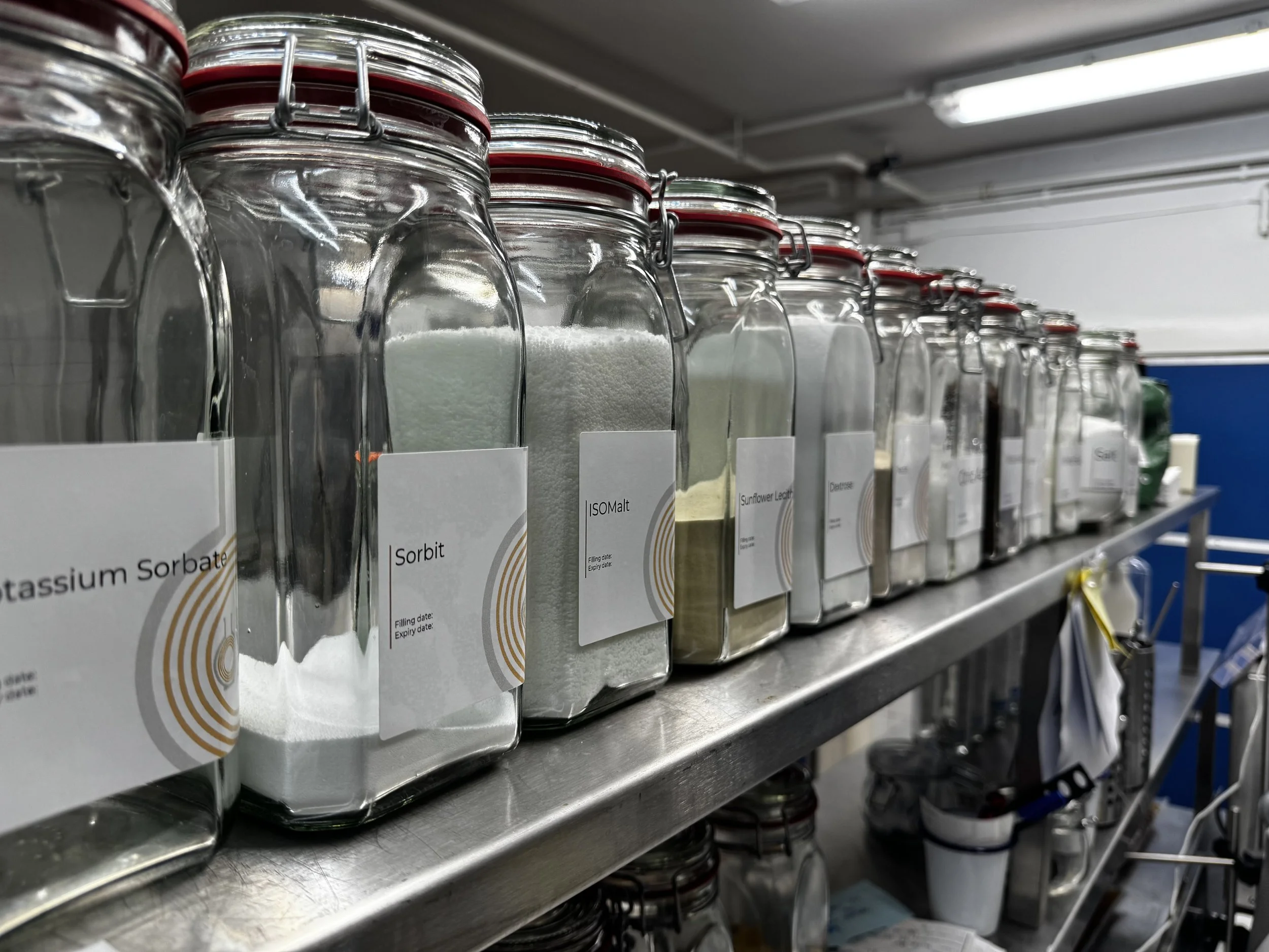 Shelf filled with glass jars containing various powdered chemicals, each labeled with their names, in a laboratory or industrial storage area.