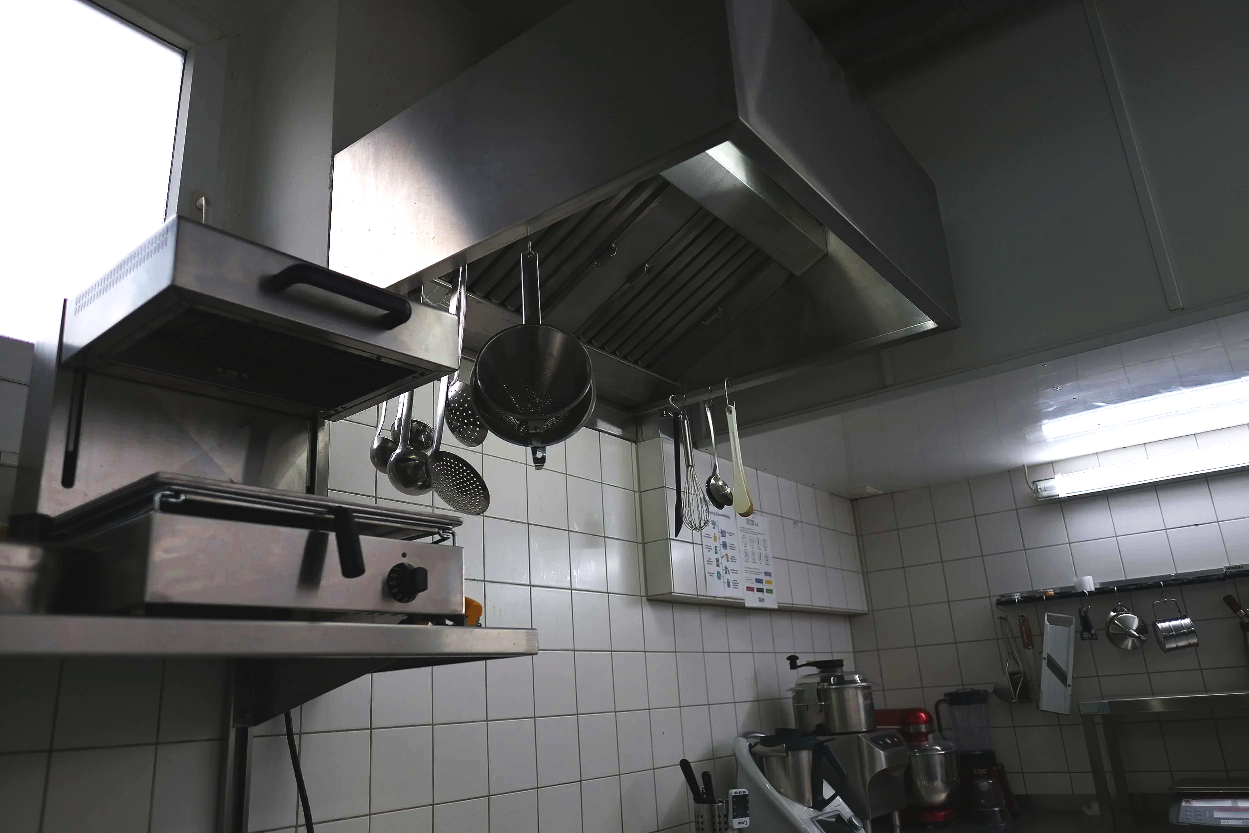 Commercial kitchen with stainless steel appliances and hanging utensils, tiled walls, and fluorescent lighting.