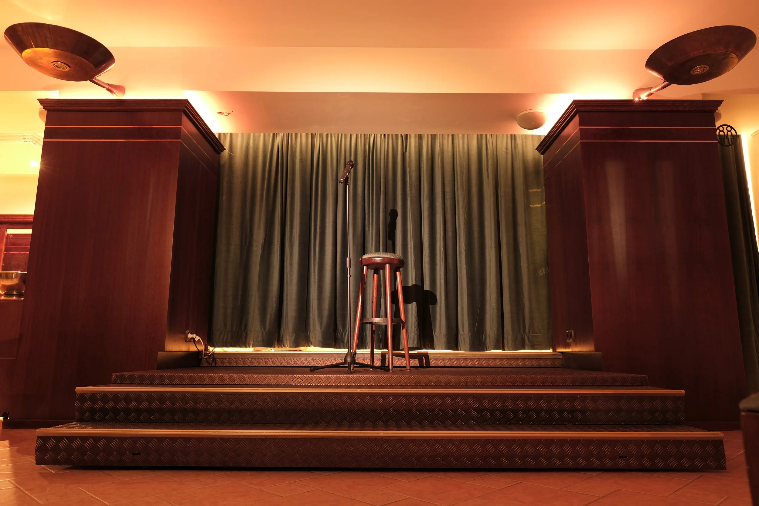 Empty comedy stage with a stool, microphone, and green curtain backdrop inside a dimly lit venue.