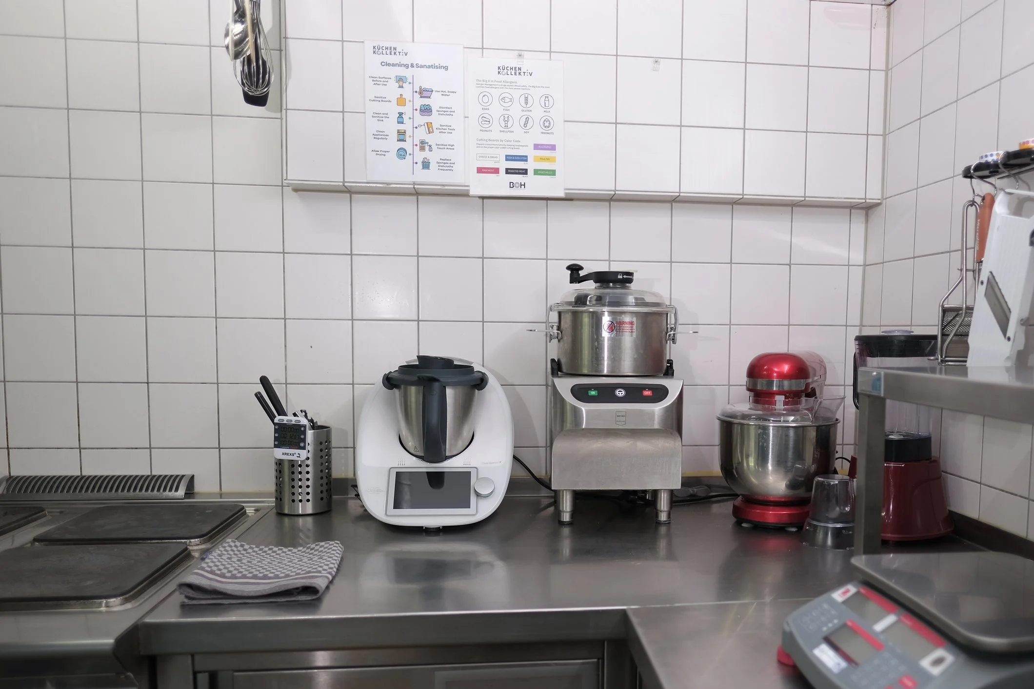 A kitchen counter with commercial appliances including a Thermomix, a robot cooker, a meat grinder, a red stand mixer, and a small food processor. There are utensils in a metal holder, a cloth towel, and a countertop calculator. The background features tiled walls and some informational posters.