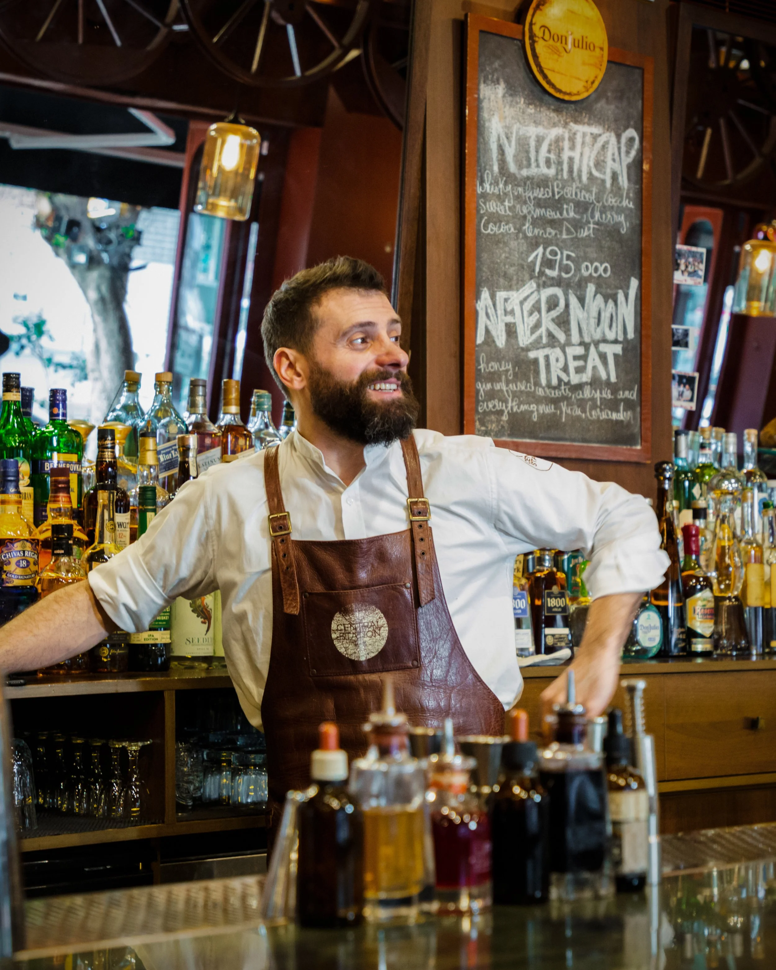 A bearded man with a white shirt and brown leather apron standing behind a bar with various liquor bottles, smiling and with arms resting on the bar counter inside a bar or restaurant.
