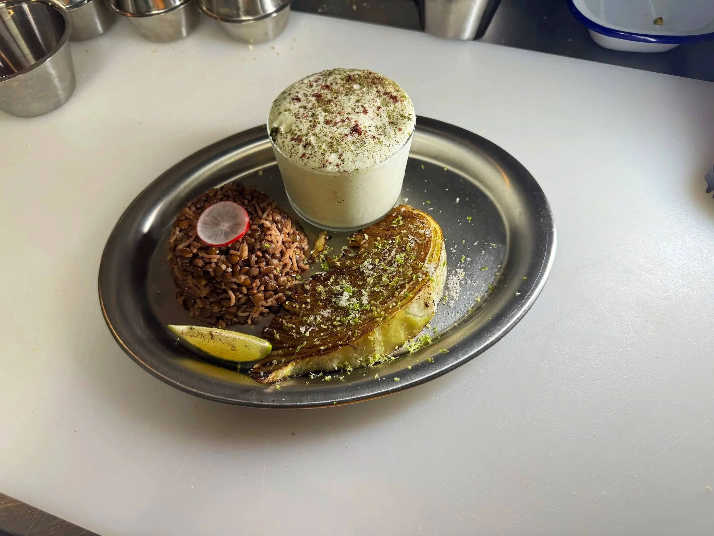 A metal plate with a serving of grilled fish, a mound of lentils garnished with a radish slice, and a glass of a creamy white dessert topped with green and red powder.