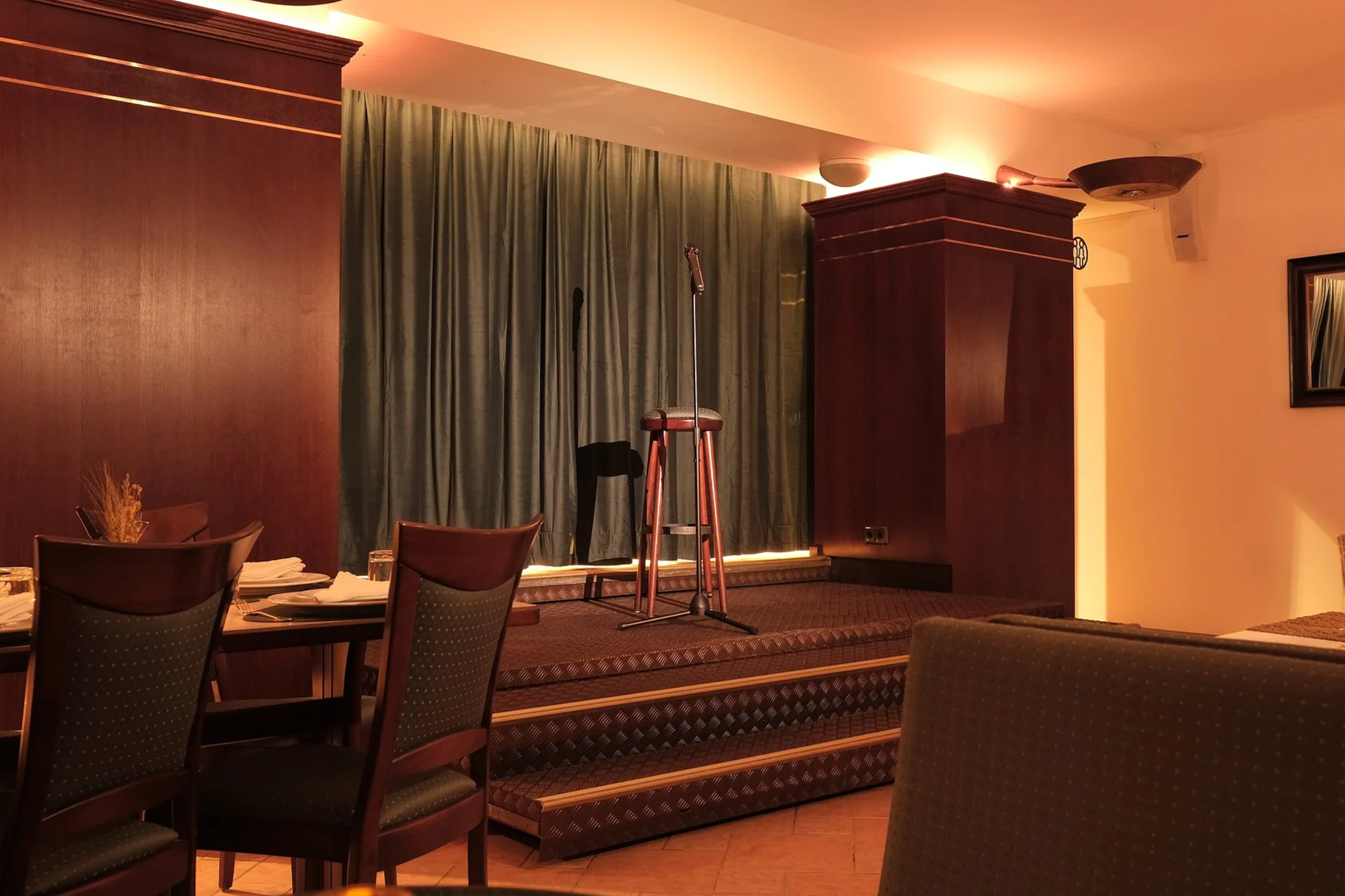 Empty stage in a restaurant with a microphone stand and stool, tables set for dining, and warm lighting