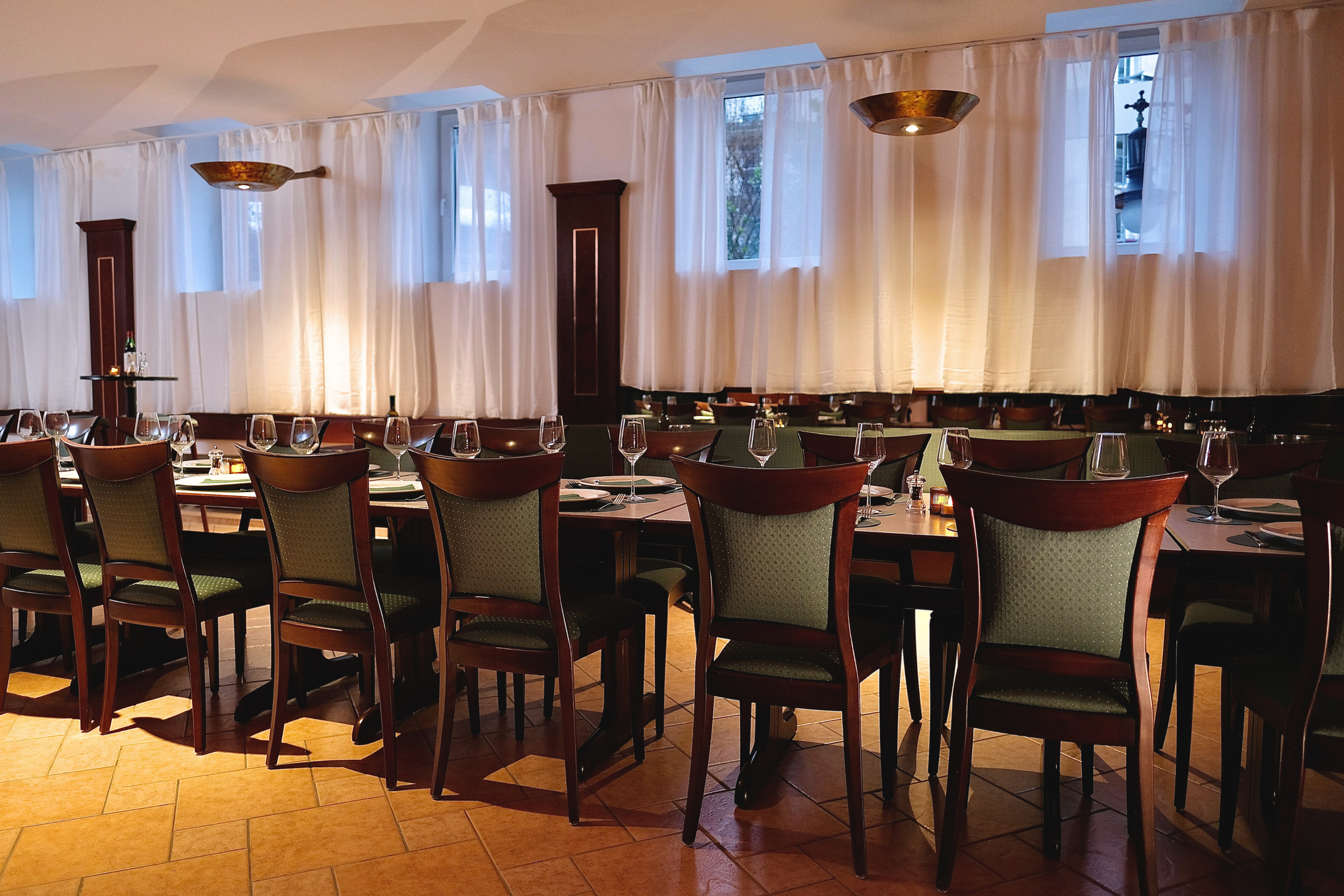 Empty restaurant dining area with wooden chairs, tables set with glasses and cutlery, large windows with white curtains, and warm lighting.
