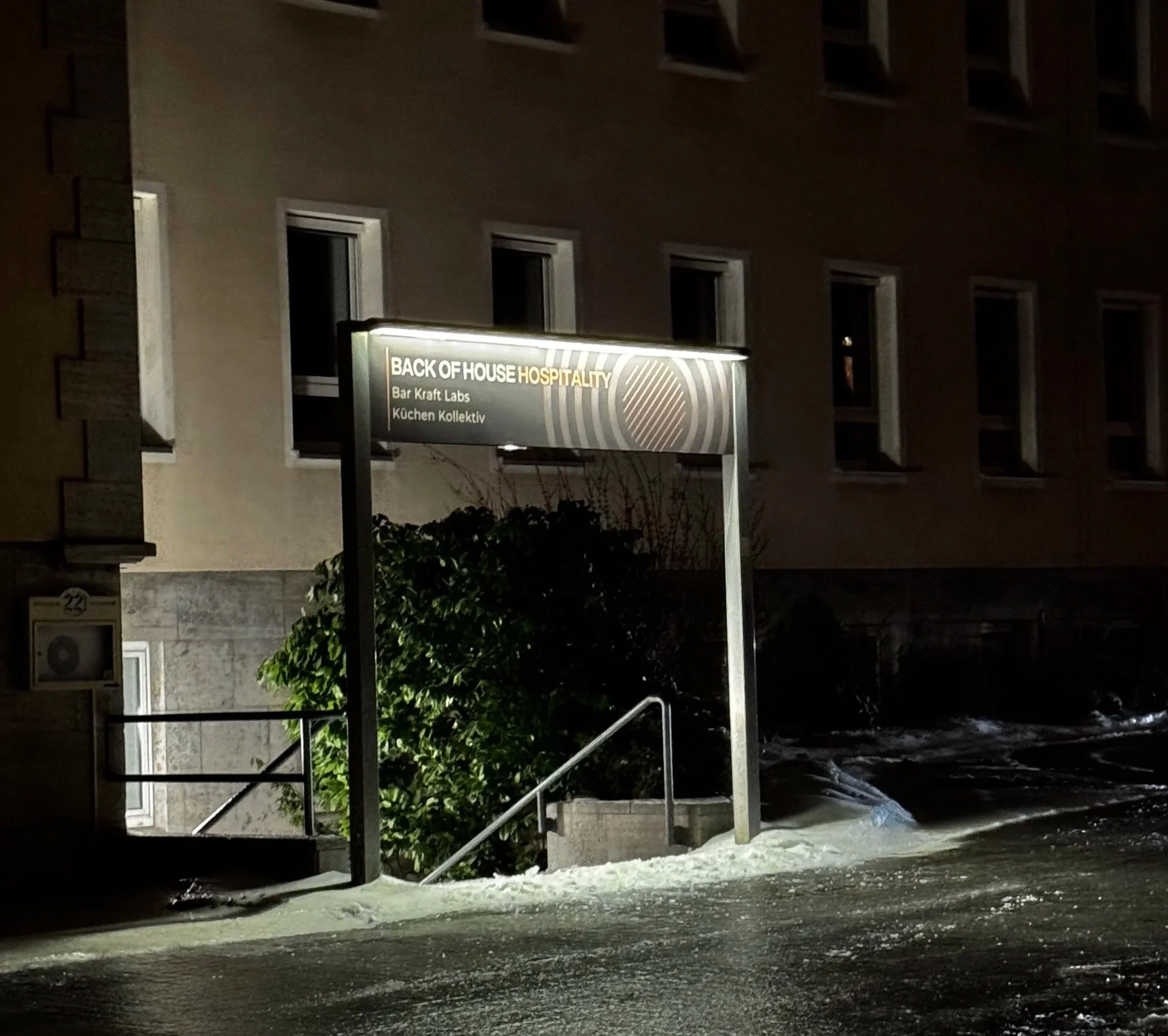 Night view of the entrance sign for 'Back of House Hospitality,' illuminated, on a street with snow and ice. The sign mentions 'Bar Kraft Labs' and 'Küchen Kollektiv.' A bush is visible below the sign, and a building with several windows is in the background.