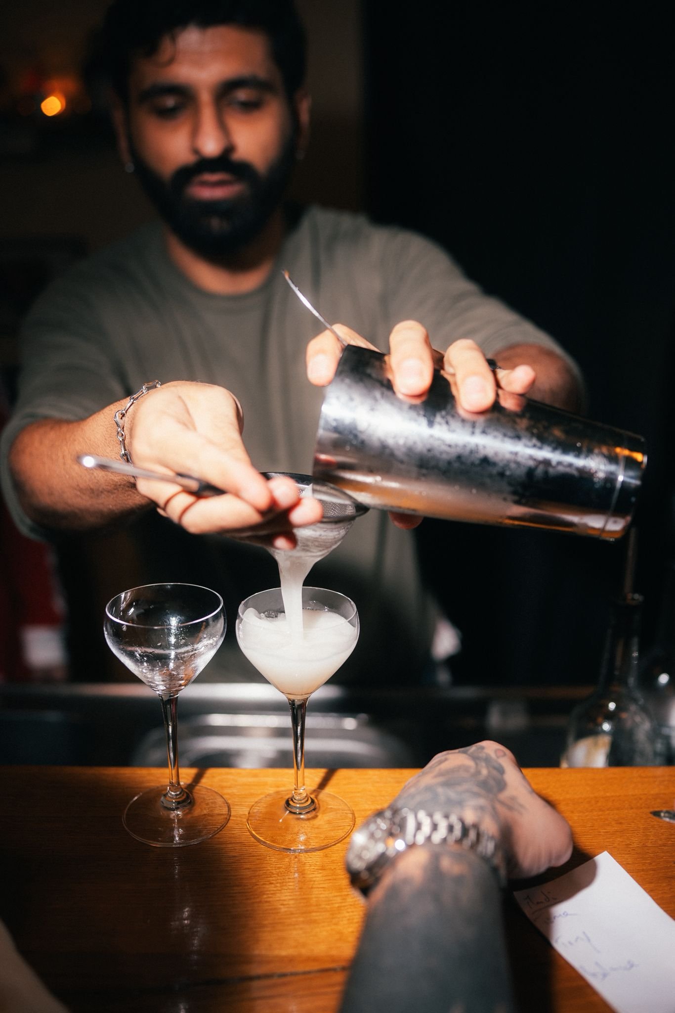 A bartender pours a drink into a cocktail glass at a bar, with two empty glasses on the wooden bar counter.