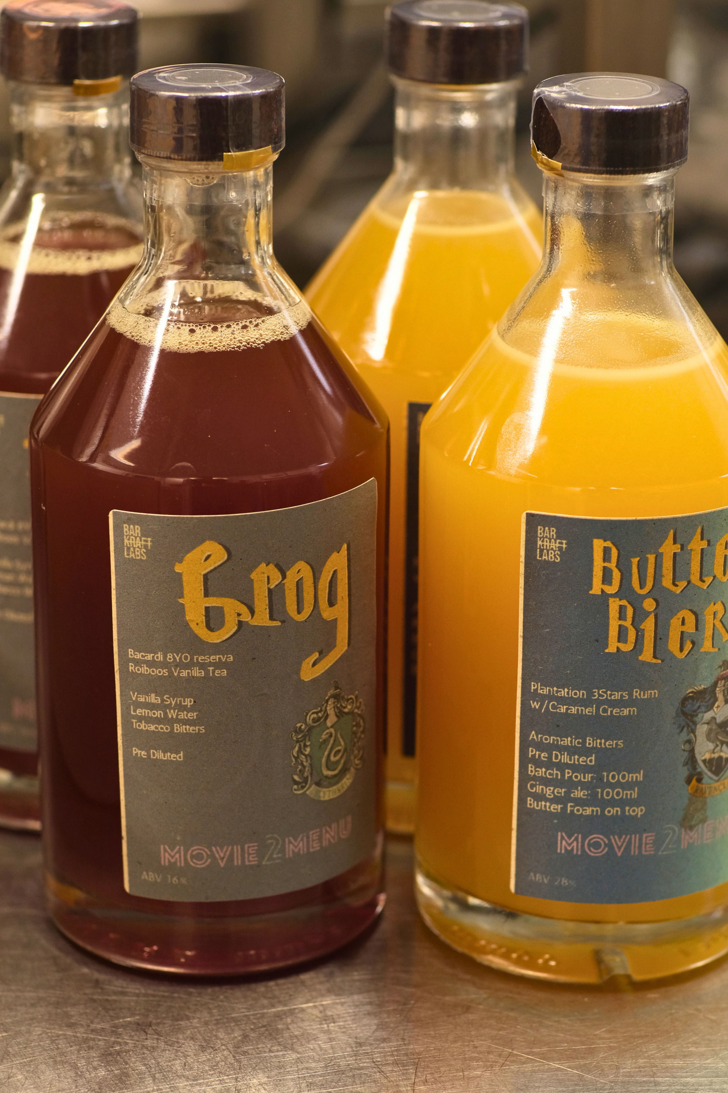 Two bottles of flavored syrup, one with a dark red liquid labeled 'Frog' and the other with a yellow liquid labeled 'Butter Bier,' on a countertop.