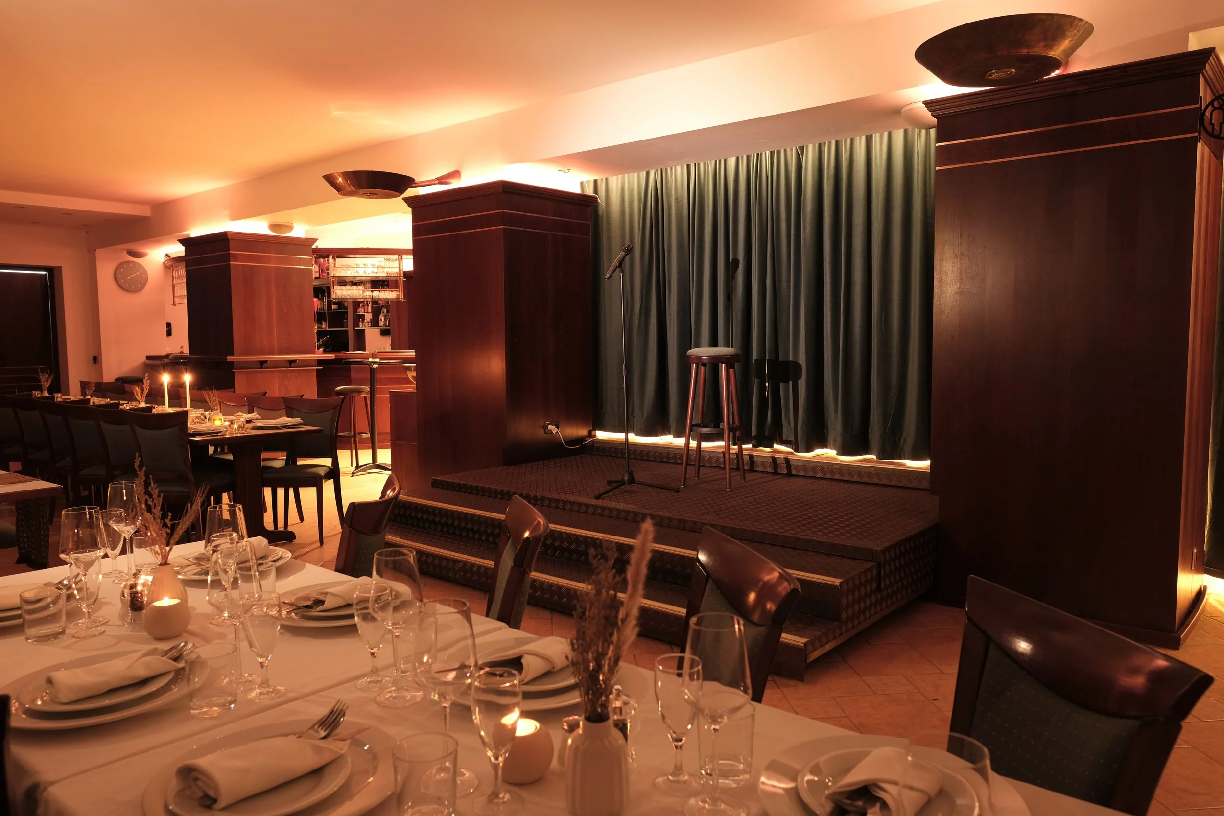 A dimly lit restaurant with a small stage featuring a microphone, stool, and dark green curtains, surrounded by elegantly set tables with white tablecloths, glasses, plates, and candles.