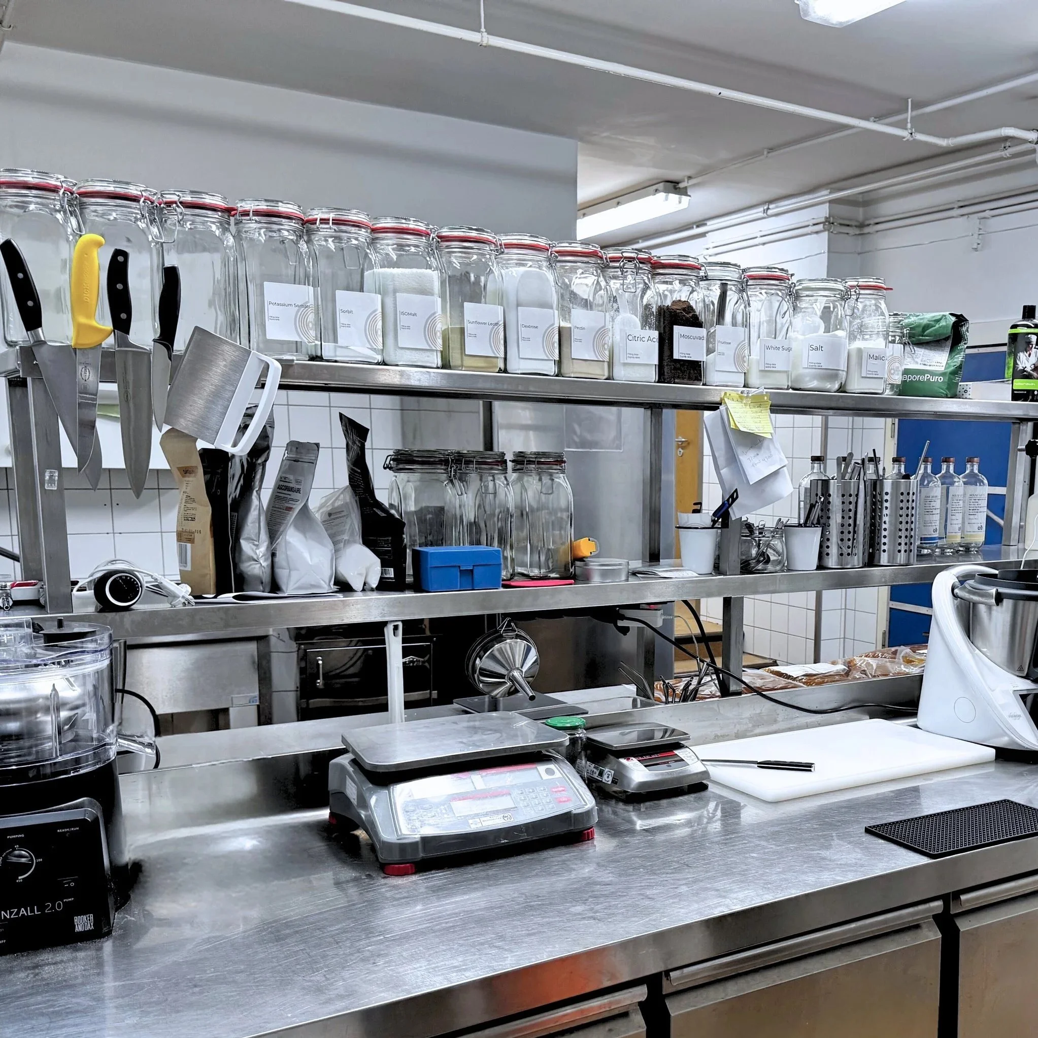 A commercial kitchen workspace with metal shelves, various kitchen tools, containers, and ingredients used for food preparation.
