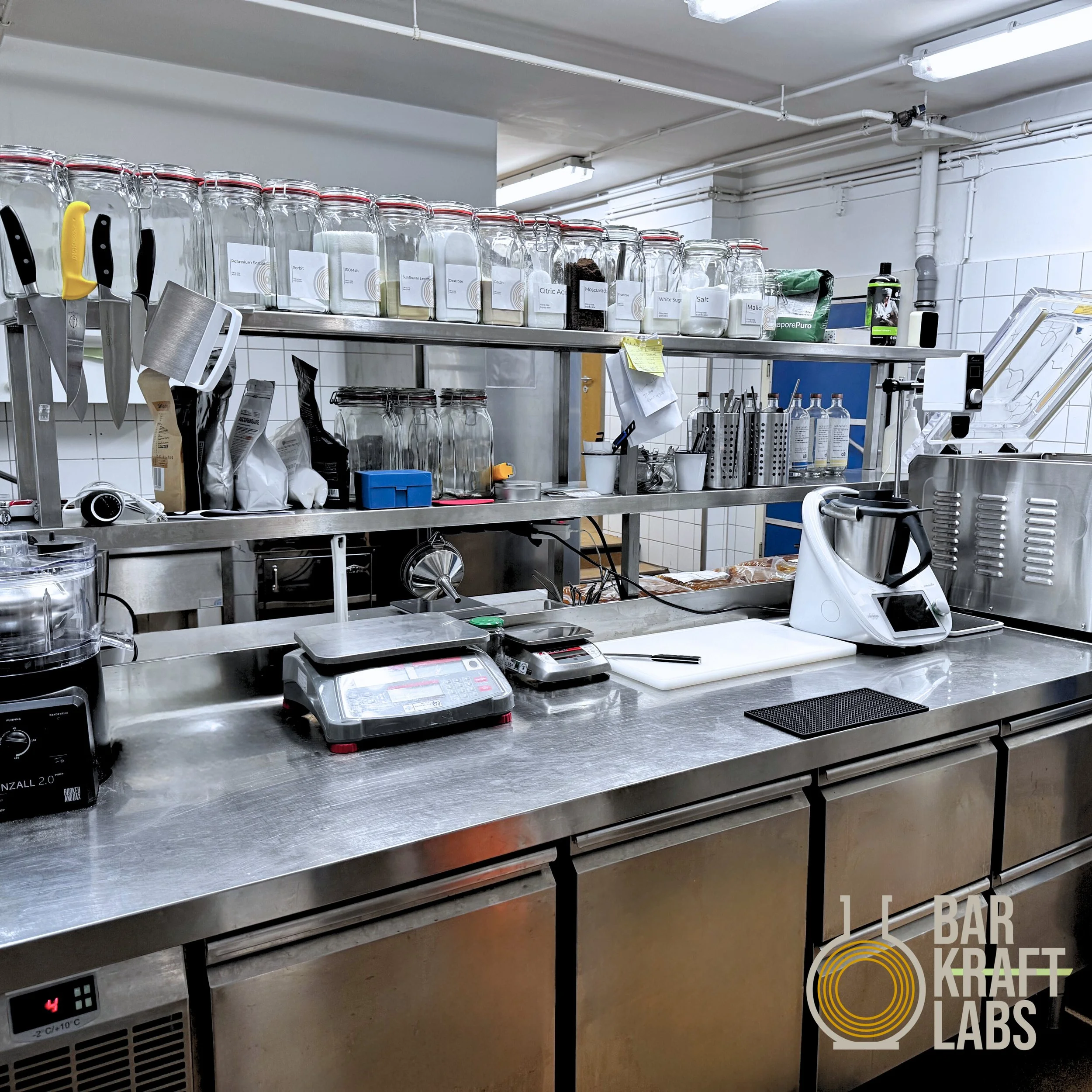 Clean laboratory or testing kitchen workspace with glass jars on shelves, kitchen tools, electronic devices, and a stainless steel countertop.
