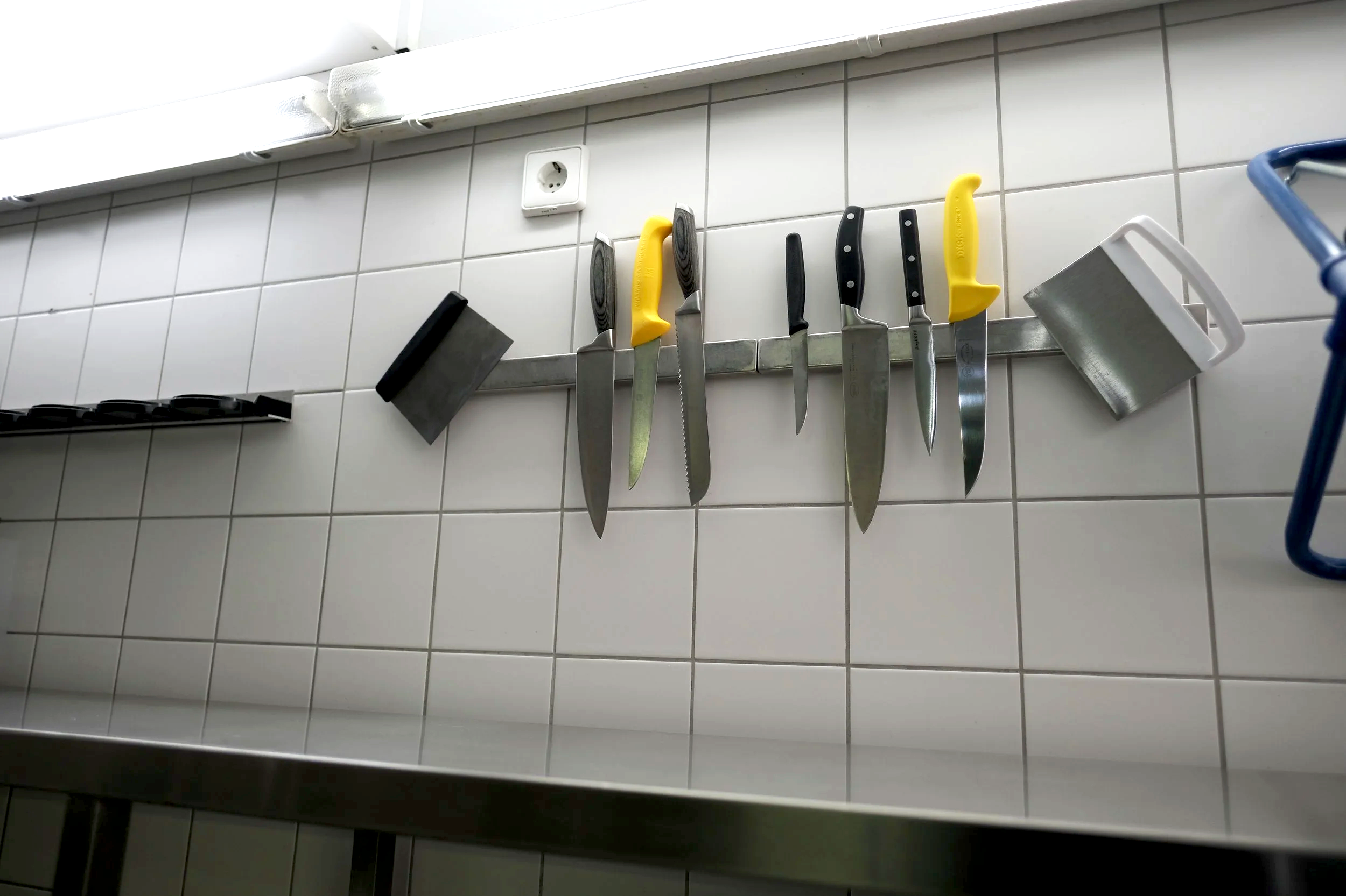 A row of knives hung on a magnetic strip mounted on a white tiled kitchen wall.