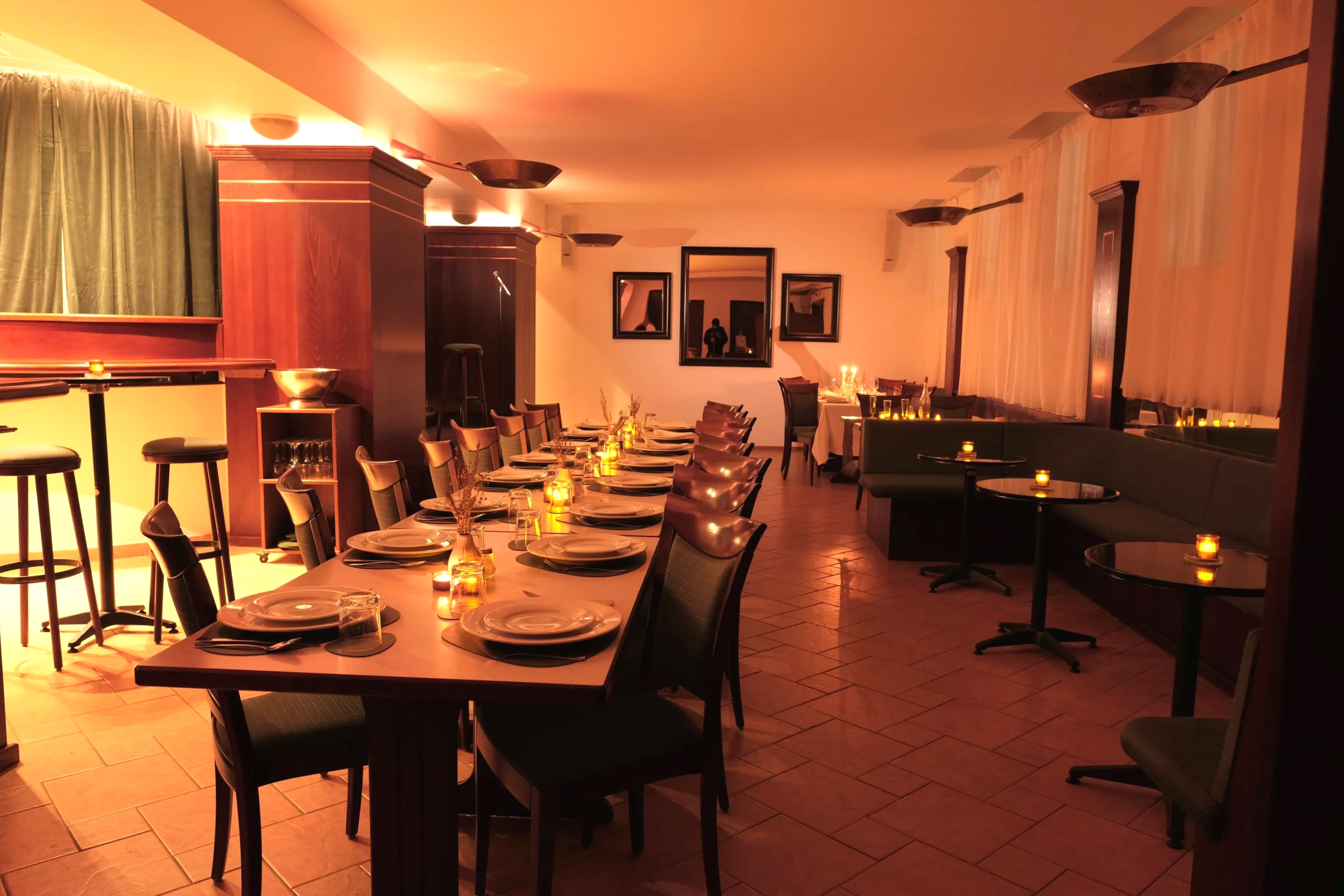 An empty restaurant or private dining room decorated with warm lighting, a long table set with plates, glasses, cutlery, and small candles, along with additional tables and chairs along the walls.